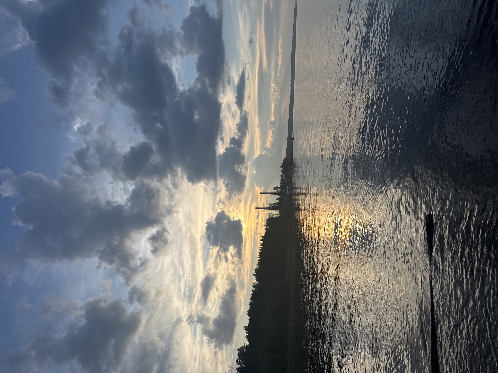 This image depicts a tranquil sunset over the Kama River in Chaikovsky, Russia. A dark, silhouetted treeline borders the left side of the frame, behind which two industrial chimneys rise. The sky is a dramatic canvas of blue, with heavy, dark clouds interspersed with patches of lighter, illuminated clouds where the setting sun is breaking through. The sun's golden light reflects off the calm surface of the river, creating a shimmering path of light. In the foreground, a portion of a dark, horizontal object, likely an oar or a log, is visible extending into the water from the left side of the image. The overall scene conveys a sense of peace and the natural beauty of the industrial city.