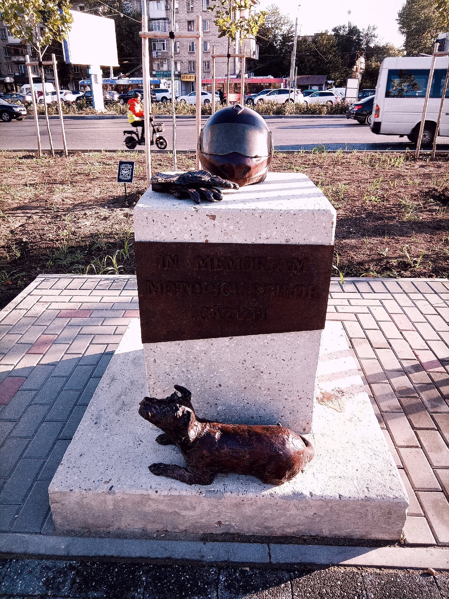 The image depicts a memorial monument located in Chișinău, Moldova. The monument is situated on a paved sidewalk with brick-like paving stones, adjacent to a street with vehicular traffic and a sidewalk lined with young trees. A white van is parked on the right side of the street. In the foreground, a concrete pedestal supports a metal plaque inscribed with text. Above the plaque, a motorcycle helmet and a pair of black gloves are placed on the pedestal. At the base of the pedestal, a bronze sculpture of a dog is positioned. The background shows buildings, trees, and a street with various vehicles, including cars and a bus. A scooter with a rider wearing a high-visibility vest is visible in the middle ground on the street. A small sign with a logo is planted in the dirt area next to the paved sidewalk. The lighting suggests it is daytime, with sunlight casting shadows.