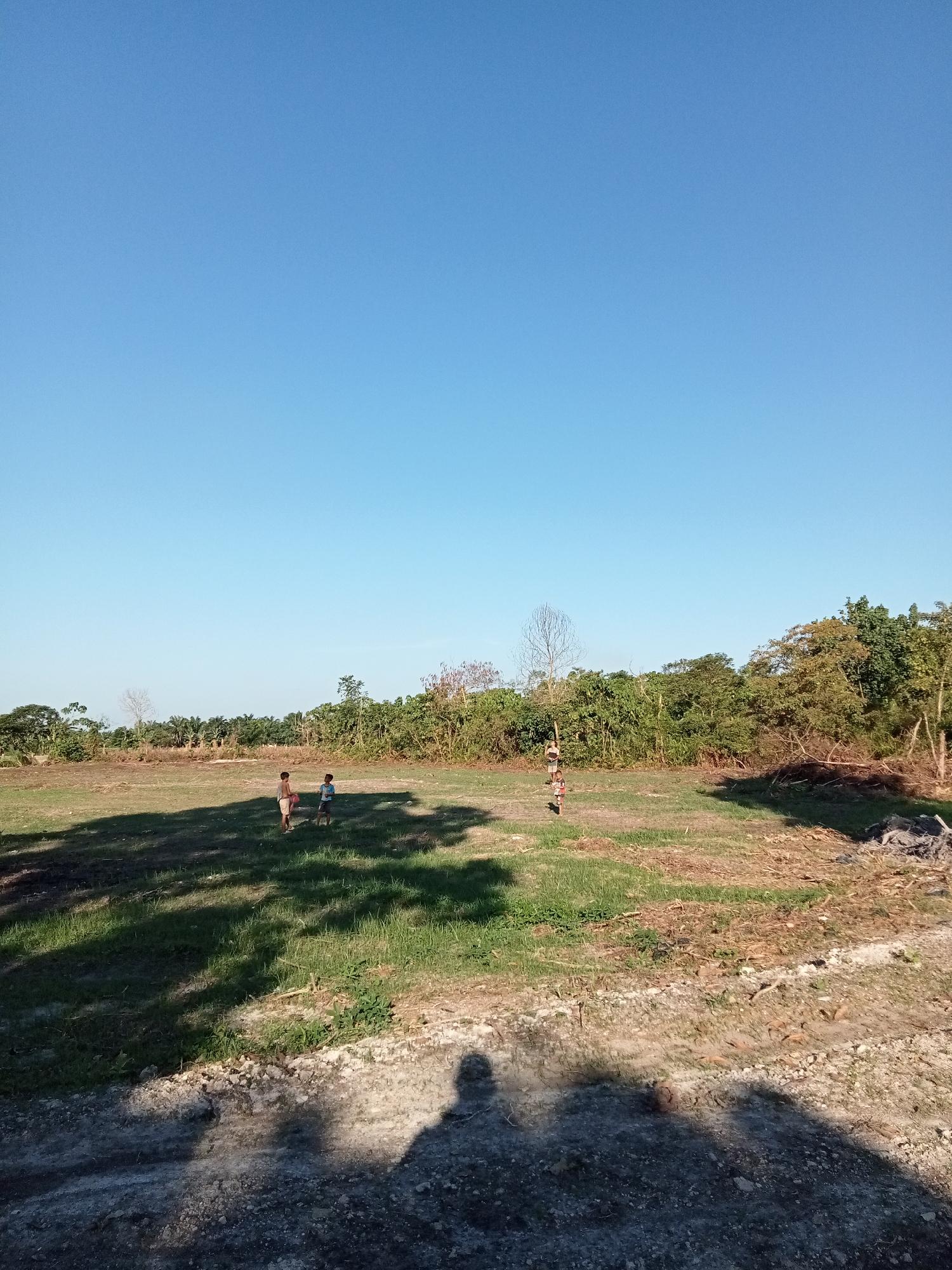 In Kualu, Indonesia, two young boys are playing in a large, open grassy field under a clear blue sky. The field has patches of dry, brown earth and is surrounded by lush green trees. The foreground shows a shadowed area, likely from the perspective of the photographer, indicating a bright sunny day. The boys appear to be standing and possibly interacting, one holding a red object. The scene suggests a rural or semi-rural environment with natural vegetation.