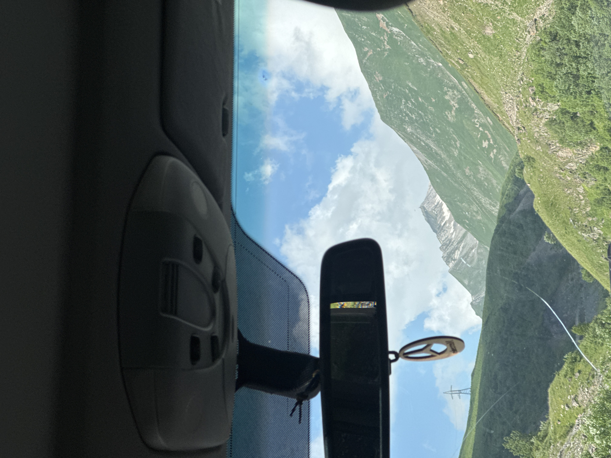 The image is taken from inside a car, looking through the windshield at a mountainous landscape. The car's interior is visible at the top, including the rearview mirror with a wooden peace sign ornament hanging from it. The windshield has a crack running across it. Outside, the scene is dominated by lush green mountains under a partly cloudy blue sky. A tall electricity pylon is visible on a distant green hillside. The area appears to be a rural or mountainous region, possibly along a road. No people or specific actions are depicted. The location is identified as Almasiani, Georgia.