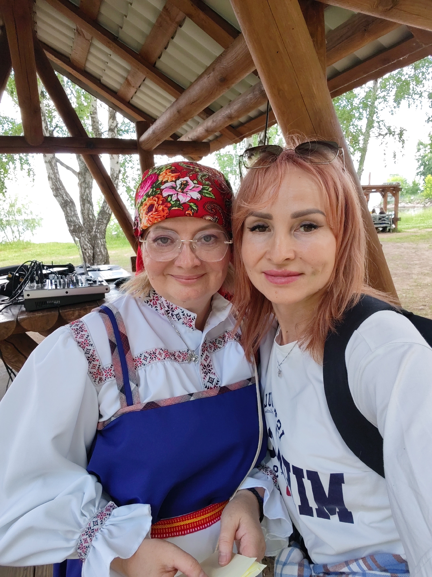 Two women are photographed outdoors under a wooden gazebo. The woman on the left wears a traditional white embroidered blouse with a blue dress over it, and a red floral headscarf. She has blonde hair, glasses, and is smiling at the camera. She is holding a small yellow object. To her left, on a wooden table, is a DJ mixer and a laptop. The woman on the right has pinkish-blonde hair and is wearing sunglasses on her head. She is wearing a white t-shirt with dark lettering and a black backpack. She is also smiling at the camera. The gazebo has a corrugated metal roof and wooden beams. In the background, there are trees and a body of water, suggesting a location near the Kama River, which flows through Chaikovsky, Russia. The overall setting appears to be a recreational area or park.