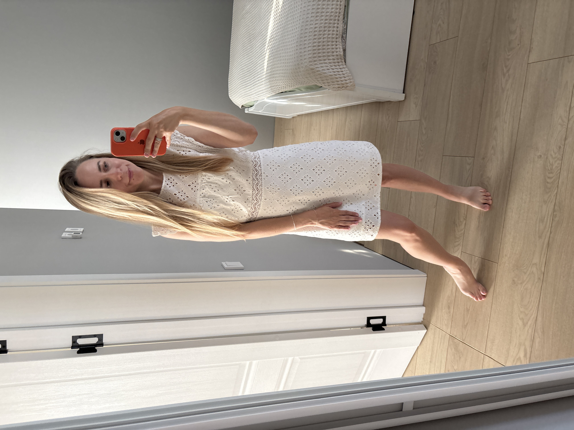 A woman stands in what appears to be a bedroom, holding an orange-cased smartphone to take a selfie. She is wearing a short white eyelet dress with cap sleeves and a lace waistband. The room has light wood flooring and a grey wall. To the left, a white door with black hardware is partially open. To the right, the edge of a white bed with a textured beige cover is visible. Sunlight casts shadows across the floor and the bed. The woman's legs are visible below the hem of her dress, and she is barefoot, with her right foot tilted. There are no other people or animals present in the image. The setting does not provide any visual cues to confirm the location as Chaikovsky, Russia.