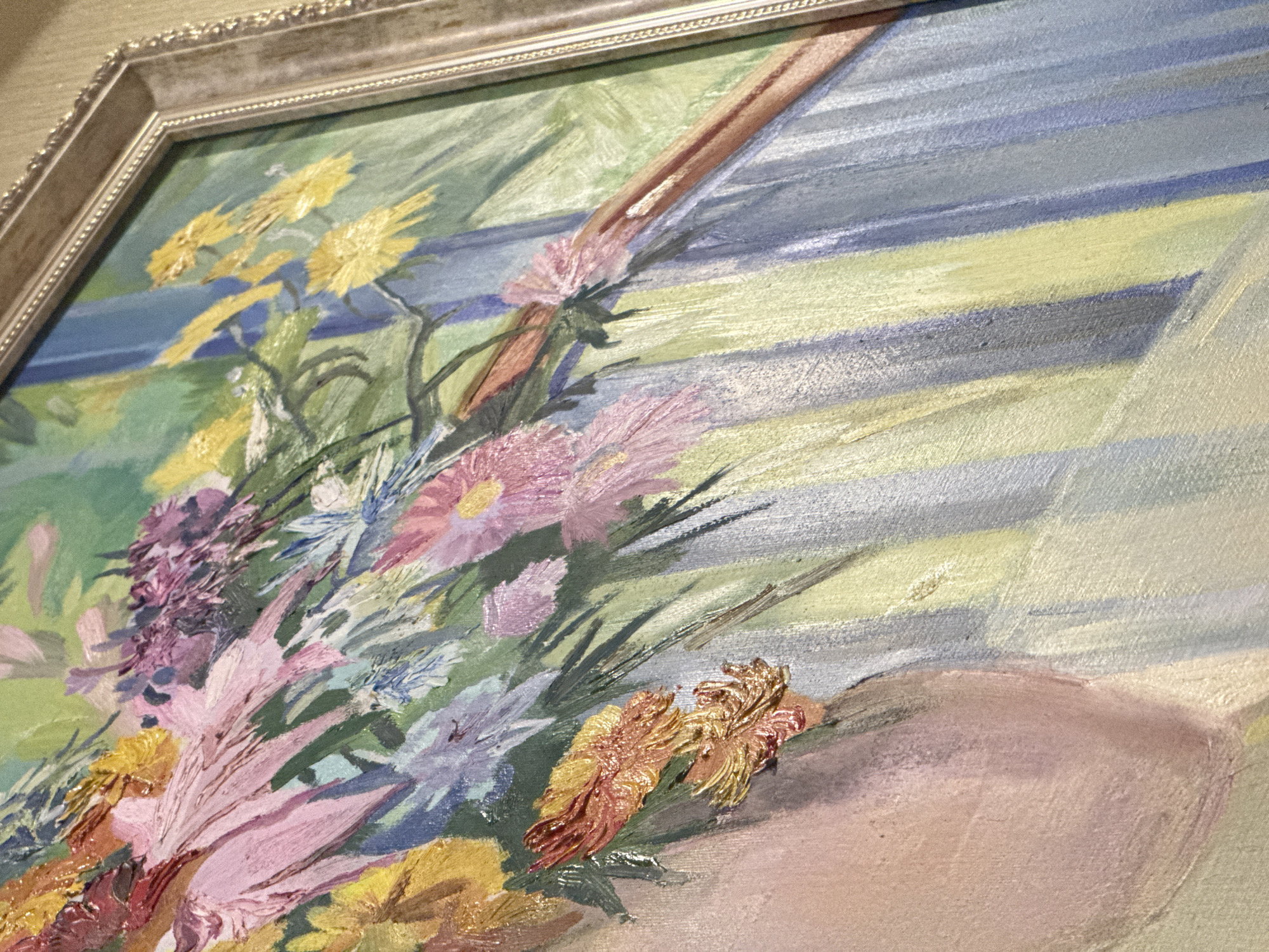 This is a close-up, angled shot of a painting of a bouquet of flowers. The painting is framed and appears to be an oil painting with visible brushstrokes and impasto texture. The flowers are a vibrant mix of pink, yellow, purple, and orange. Behind the flowers, there are vertical stripes of blue, green, and yellow, suggesting a background or a draped fabric. A portion of what appears to be a vase or the shoulder of a figure is visible on the left side of the frame, rendered in soft pink and peachy tones. The painting is displayed on a wall with a textured, light brown finish. There is no information or visual cue within the image that places it in Vinnytsia, Ukraine, or any specific city.