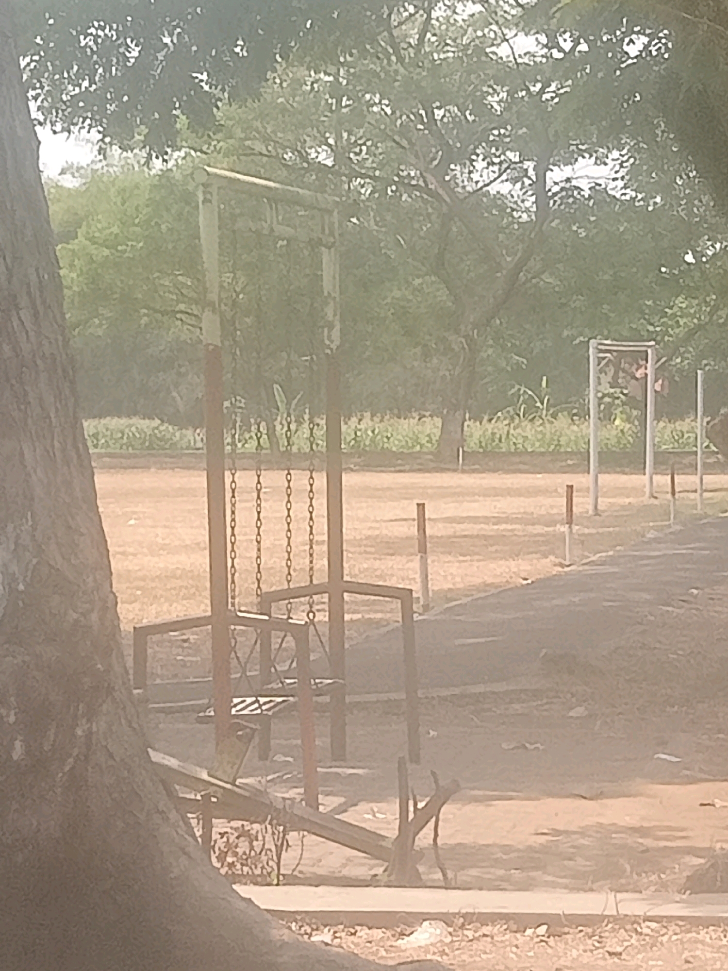 This is an image of an outdoor area with playground equipment. The image appears to be taken in Kambangan, Indonesia. In the foreground, a large tree trunk is visible on the left. A seesaw is partially obscured by the tree trunk and appears to be made of metal. In the mid-ground, a swing set with chains is visible. The swing set is red and weathered. Beyond the swing set, there is a sandy or dirt ground area, and further back, there are trees and foliage. In the background, a white structure that resembles a goalpost or frame is visible, along with several smaller posts. The overall impression is that of an abandoned or disused playground.