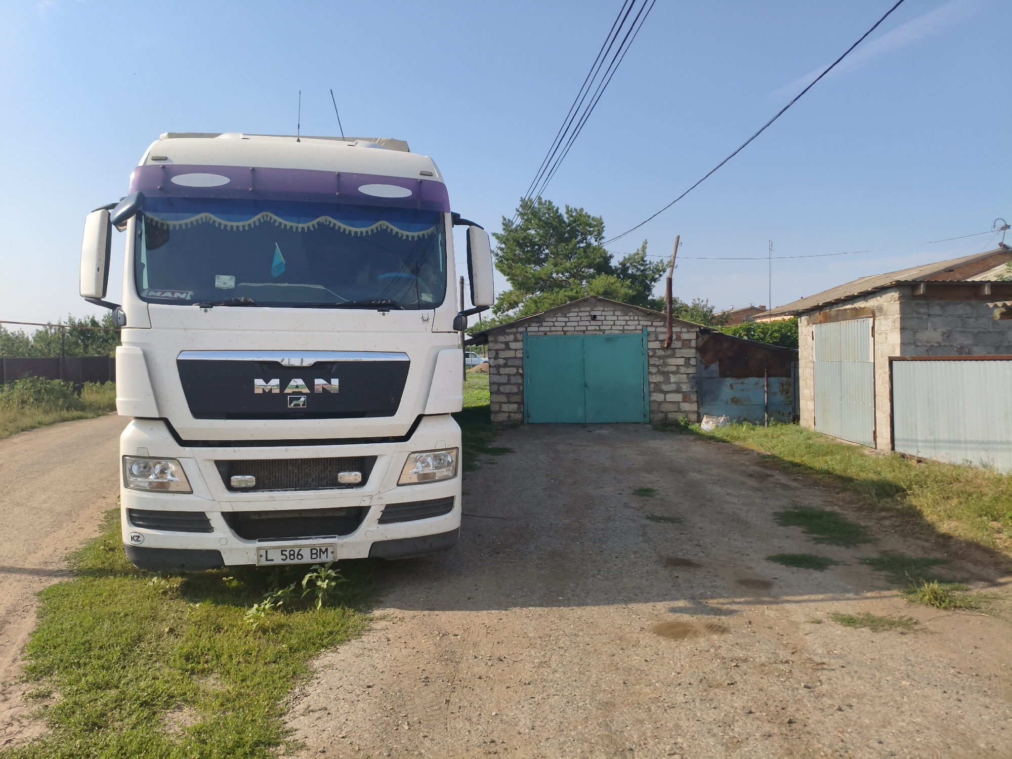 A white MAN truck is parked on a dirt road in front of a brick garage with a teal door. The truck has a license plate that reads "L 586 BM". Behind the garage, there are more buildings and trees. The sky is blue and clear, with some power lines running across it. The scene appears to be in a rural area. The location is Ақсуат ауылдық округі, Kazakhstan.