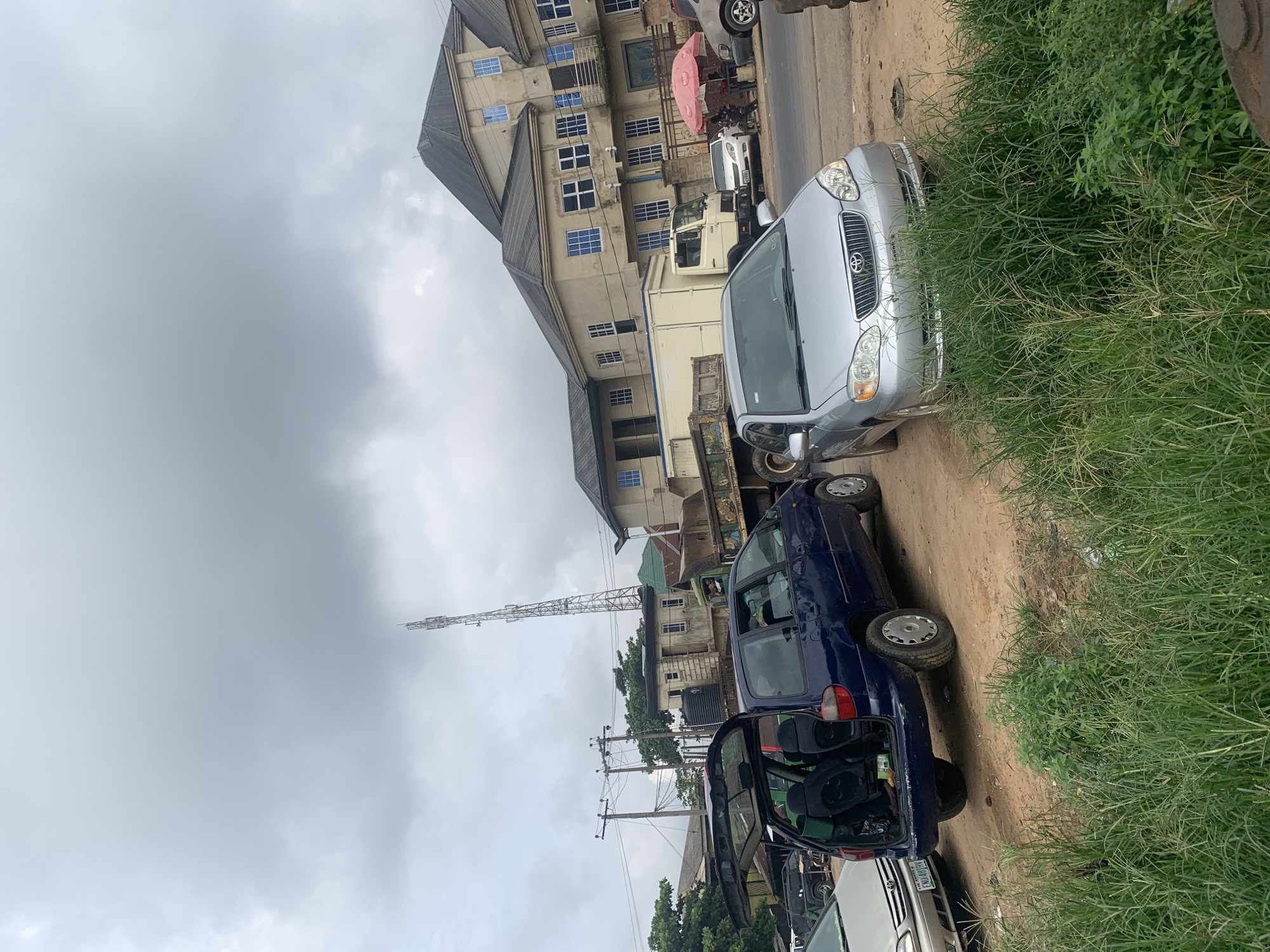 In Mbara Akuma, Nigeria, a scene unfolds with a cluster of vehicles and buildings under an overcast sky. A silver Toyota sedan is parked in the foreground, facing right, its hood and headlights visible. Behind it, a dark blue minivan with its rear hatch open reveals a glimpse of its interior. A white box truck is positioned further back, partially obscured by the minivan. A large, industrial-looking truck with a weathered bed sits to the right of the box truck. In the background, a tall cell tower rises into the cloudy sky. Several multi-story buildings with light-colored facades and blue-tinted windows line the street. A red umbrella offers shade over a small group of people gathered near a table by the roadside. The ground is a mix of dirt and sparse vegetation.