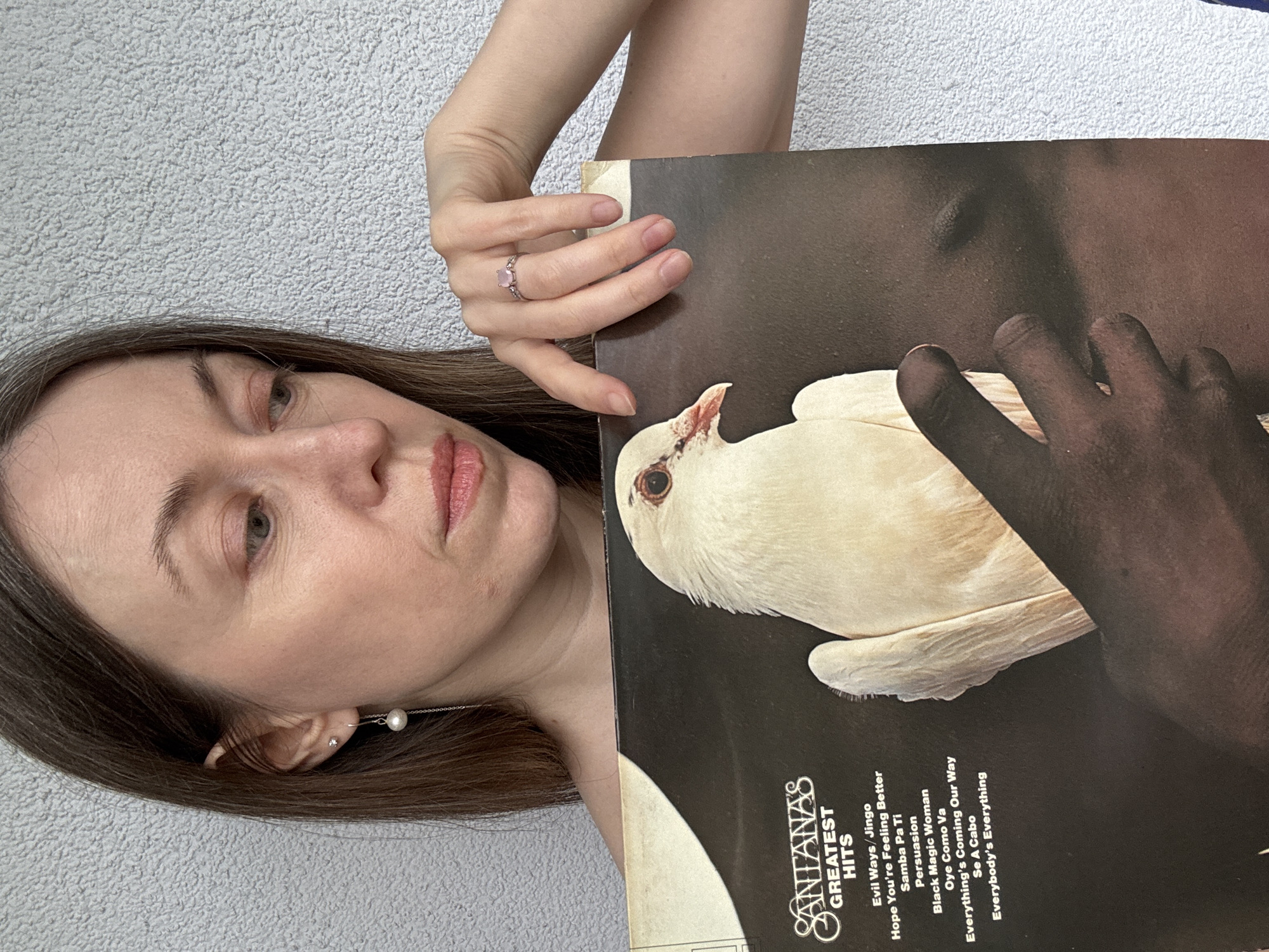 A medium shot captures a woman holding up a vinyl record album against a textured, light gray wall, likely in Kudrovo, Russia. The woman has long brown hair, fair skin, and light-colored eyes, looking slightly to her left with a neutral or pensive expression. She wears a silver ring with a pink heart-shaped stone on her right ring finger and a delicate silver earring with a pearl drop in her left ear.
She is holding the "Santana's Greatest Hits" album. The album cover art features a dark background with a dark-skinned hand cradling a white dove.
Visible text on the album cover includes "SANTANAS GREATEST HITS" and a tracklist: "Evil Ways/Jingo", "Hope You're Feeling Better", "Samba Pa Ti", "Persuasion", "Black Magic Woman", "Oye Como Va", "Everything's Coming Our Way", "Se A Cabo", and "Everybody's Everything". The image is oriented with the woman's head tilted, appearing sideways to the viewer.