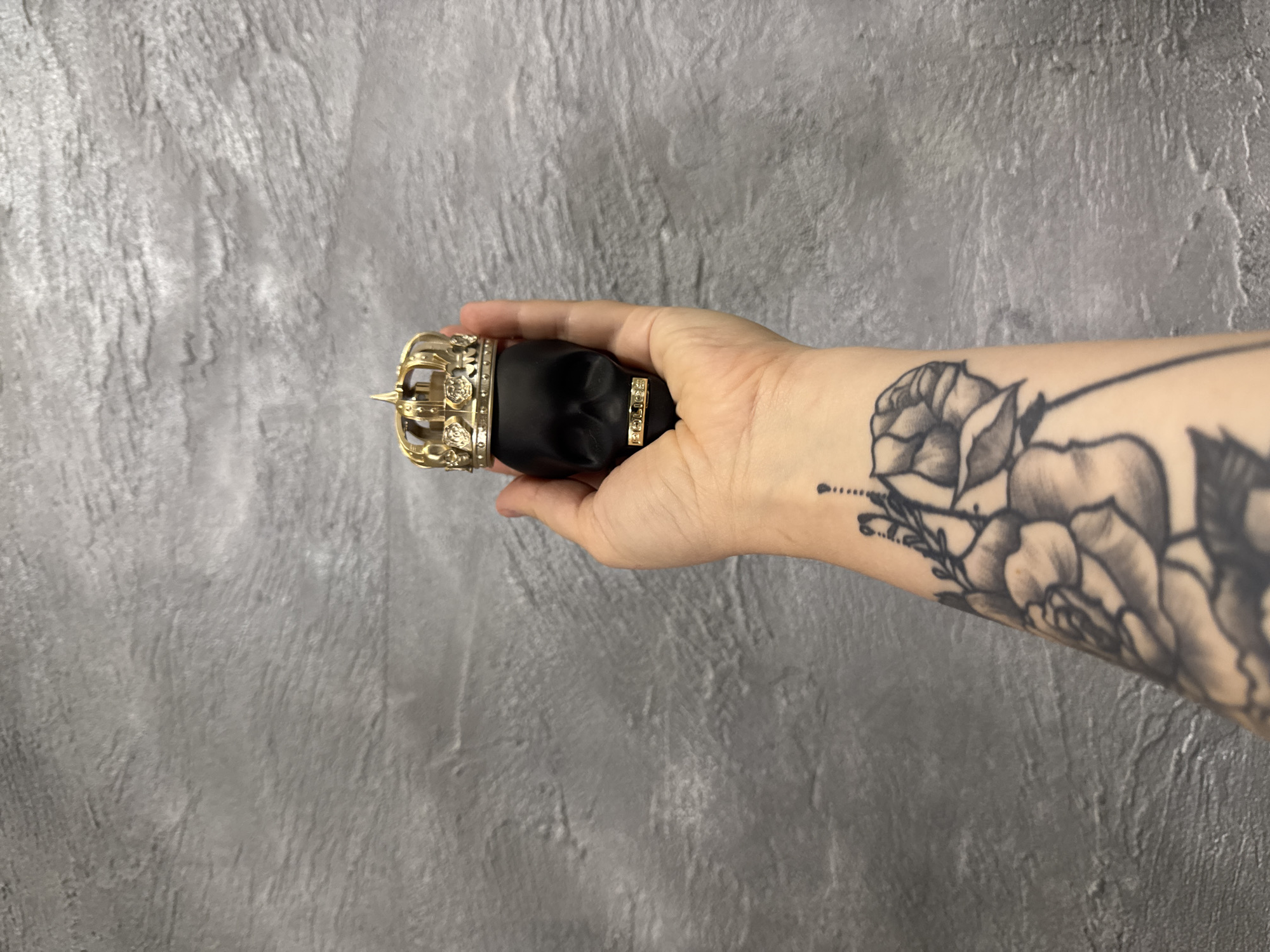 The image displays a person's fair-skinned hand and forearm holding a black, skull-shaped object against a textured grey wall. The object, possibly a perfume bottle, features an elaborate golden crown with intricate floral details (resembling roses) on its top. A golden band wraps around the skull's forehead, clearly displaying the word "POLICE" in small gold letters. The person's forearm is extensively tattooed with a black and grey floral design, predominantly featuring roses and accompanying foliage. The grey wall background has a rough, uneven texture, suggesting concrete or a similar material. The lighting appears soft and uniform, creating a clear view of the subjects. No specific time of day, weather, or location cues are apparent, and no emotions are discernible from the visible hand and arm.