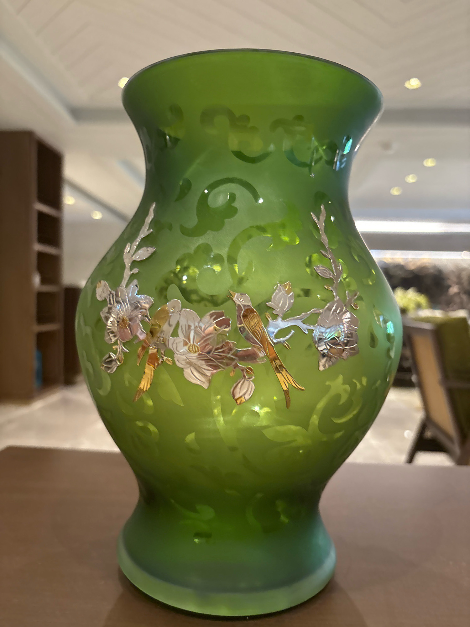 A medium shot captures a vibrant green glass vase with delicate silver and gold floral and bird embellishments. The vase is ornately decorated with swirling patterns etched into its surface, creating a frosted effect. The background is softly blurred, showing hints of a hotel lobby with muted lighting, wooden furniture, and possibly a shelf unit. The vase sits on a polished wooden surface, suggesting an indoor setting. The lighting emphasizes the translucent quality of the glass and highlights the metallic accents of the artwork. The image is well-composed, focusing on the decorative details of the vase.