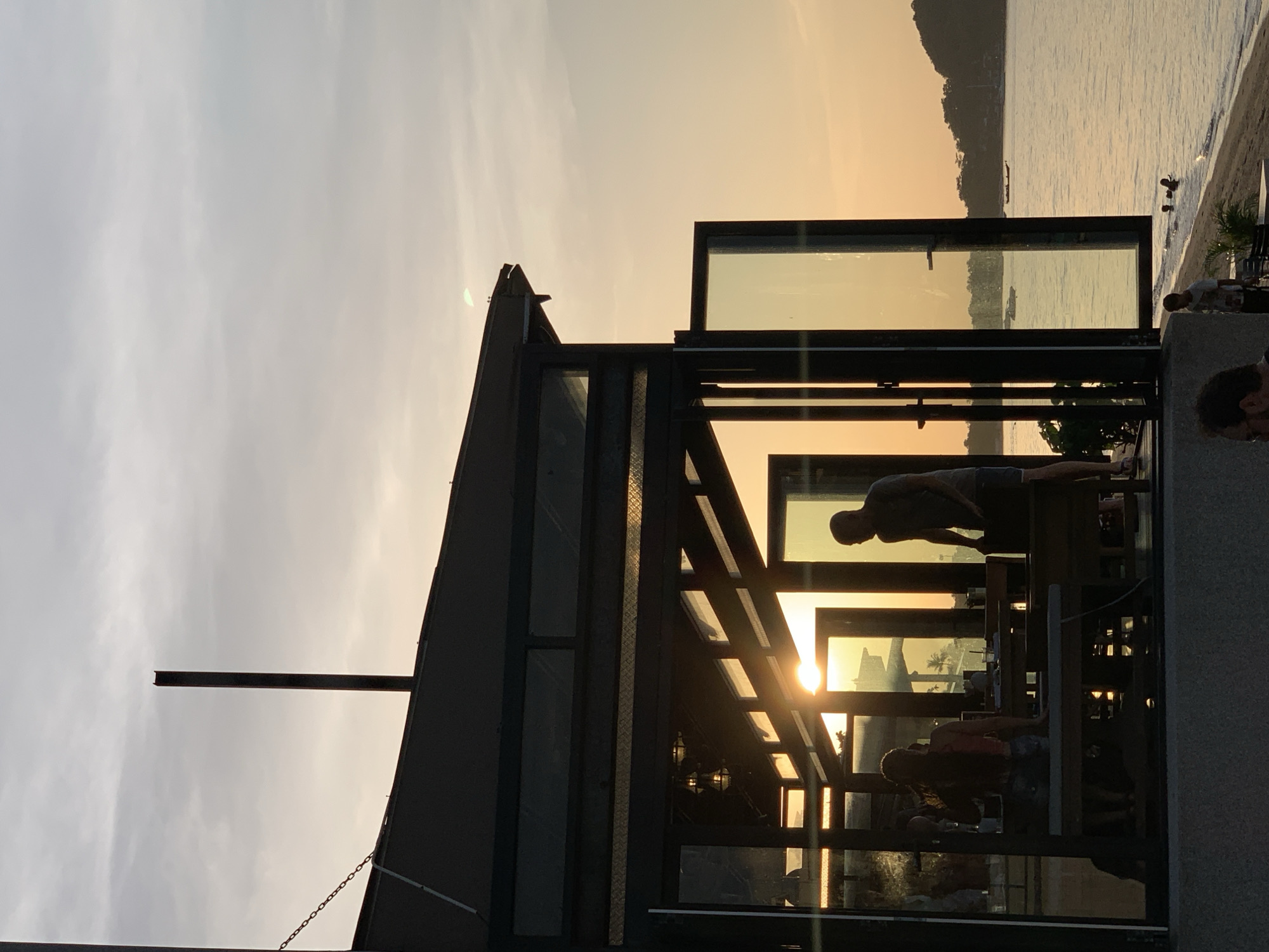 A silhouetted building with large glass panels and an open section stands on the coast in Ko Samui District, Thailand, during sunset. Inside the building, two people are visible sitting at tables, with the setting sun creating a strong backlight that obscures details. Outside, the sea is calm with a few boats on the horizon. A sandy beach runs alongside the water, and a few people are visible in the water near the shore. The sky is a soft gradient of orange and grey clouds. The scene suggests a coastal restaurant or bar setting at dusk.