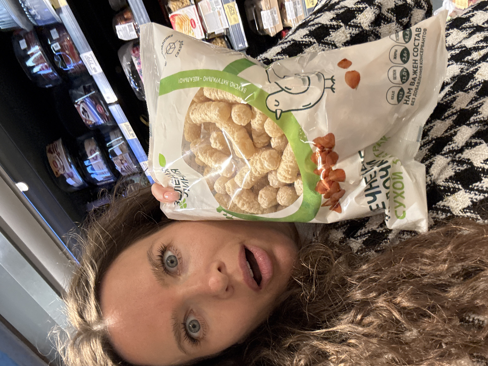 A woman with long, curly brown hair is taking a selfie in a grocery store aisle. She is holding a bag of puffed snacks made from corn or similar grain, with a cartoon bird and text in Russian on the packaging. The snacks appear to be round or elongated and light brown in color. The woman has wide blue eyes and her mouth is open in a surprised expression. She is wearing a black and white patterned jacket. In the background, shelves are stocked with various food items, including packaged cakes and other snacks. The lighting suggests an indoor commercial environment.