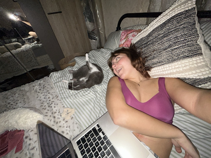 A young woman lies on a striped bedspread, smiling and looking towards a grey and white cat grooming itself with its leg raised in the air. She is wearing a purple top and has a laptop open in front of her. A large patterned pillow rests behind her head. A mirror reflects the bed and room, adding depth to the scene. Another laptop is next to her on the left.
