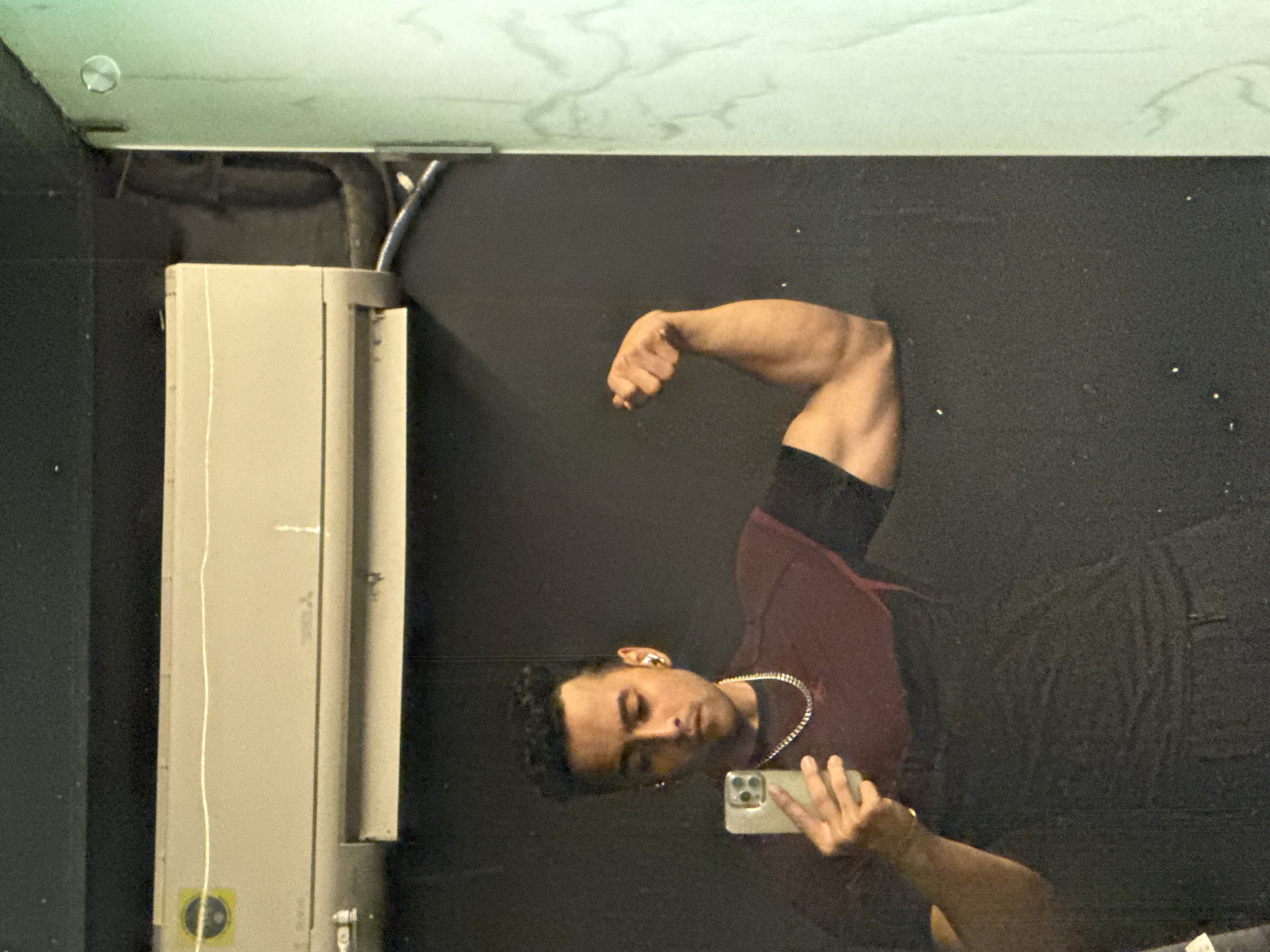The image shows a person taking a selfie in a mirrored room. The person is flexing their right bicep while holding a smartphone. They are wearing a maroon shirt, black undershirt, and a silver chain. An electrical unit is visible on the left side of the image. The background appears to be a dark wall. There is no information to suggest the location is Kolkata, India.