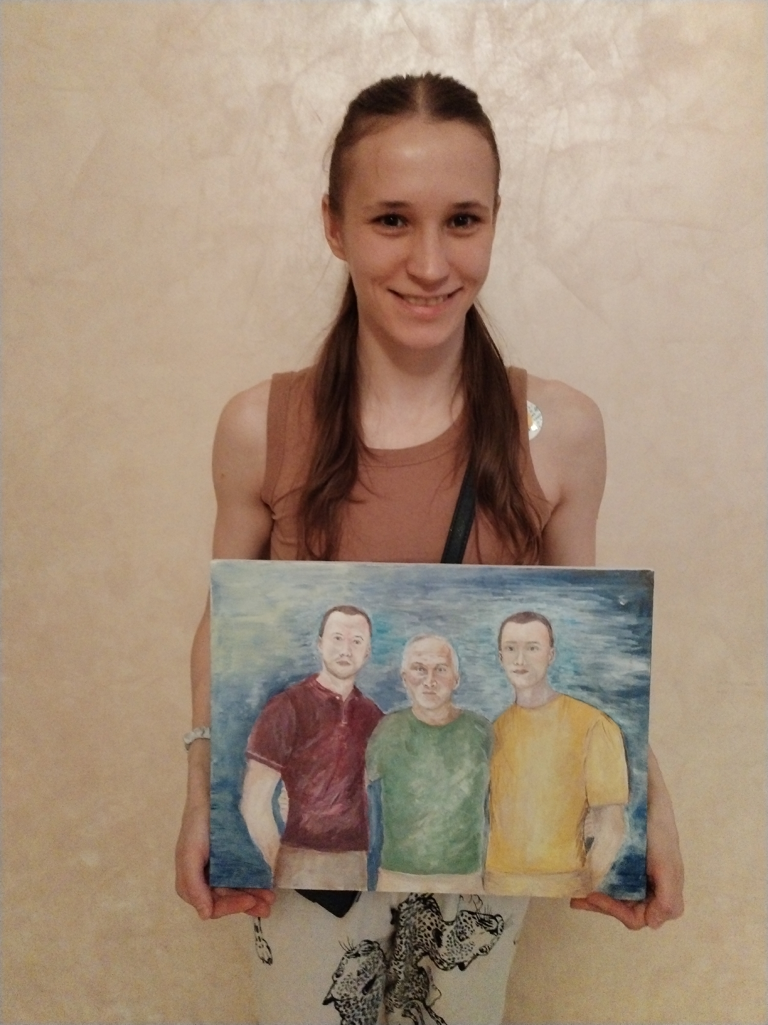 A young woman with long, dark hair styled in pigtails is the central figure, smiling brightly at the camera. She wears a light brown sleeveless top and patterned white trousers, partially visible. A black strap is slung over her left shoulder, and a small, silver, round patch is visible on her right upper arm.
She is holding a rectangular painting with both hands, positioned in front of her torso. The painting depicts three men standing side-by-side against a blue and white abstract background. From left to right, the painted men wear a red, green, and yellow short-sleeved shirt respectively. The central painted figure appears older with light hair, flanked by two younger men with dark hair. Their expressions are neutral.
The woman is posing for the camera, presenting the artwork, conveying a sense of pride or happiness. The background is a plain, light-colored, textured wall, indicating an indoor setting. No visible text appears in the image. This personal moment takes place indoors, likely in Chaikovsky, Russia.