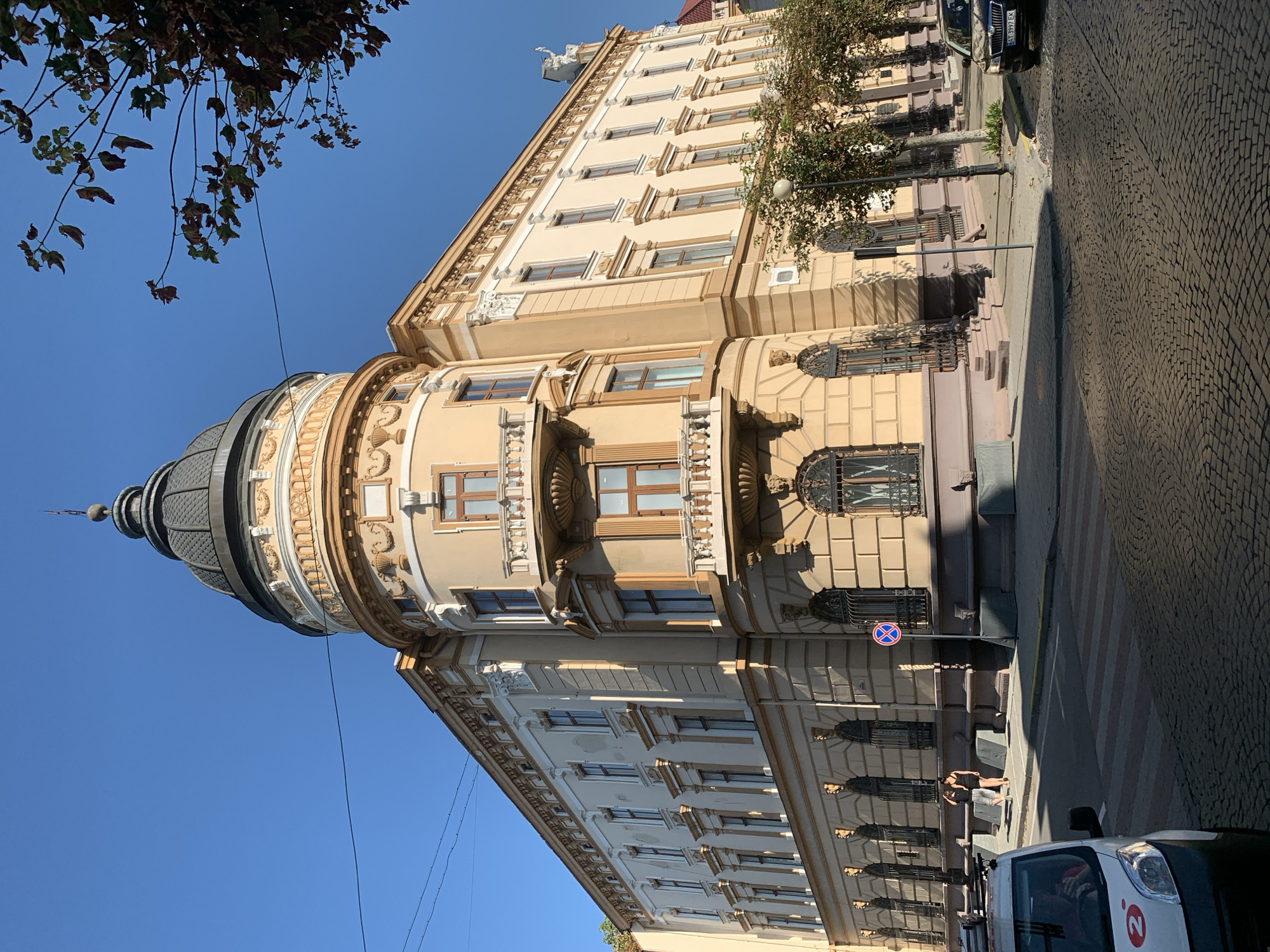 The image shows a corner of a historic building in Kolomyia, Ukraine, with a prominent dome and ornate architectural details. The building is painted in shades of beige and ochre, with white decorative elements around the windows and along the roofline. A rounded turret with a dark dome and spire rises from the corner. Multiple windows, some with decorative grilles and balconies, are visible. The building is situated on a cobblestone street. A pedestrian crossing is marked on the street in front of the building. In the foreground, the front of a white van with a red logo is visible. To the right, a dark-colored car with a license plate is parked. Two women are standing on the sidewalk in front of the building, near a street sign. Trees with sparse foliage line the sidewalk. The sky is clear and blue. The scene appears to be set during daylight hours, with sunlight casting shadows.