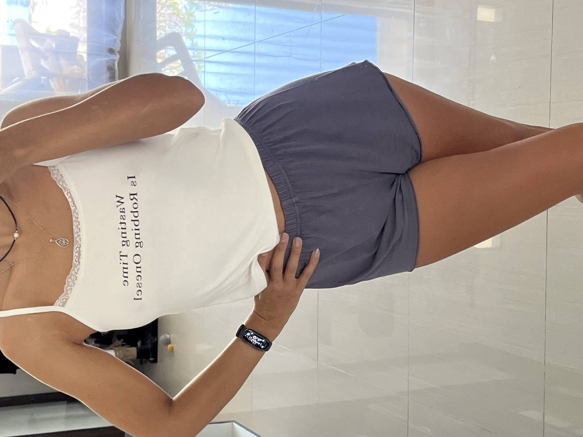 A woman is standing indoors. She is wearing a white tank top with lace trim and the words "Waiting Time Is Running Out" printed upside down. She is also wearing grey shorts and a black fitness tracker on her wrist. Her right hand is placed near her right hip. She is wearing necklaces. The background shows a window with a view of a building or outdoor area. The location is Unknown, Indonesia. The floor is light-colored tile.