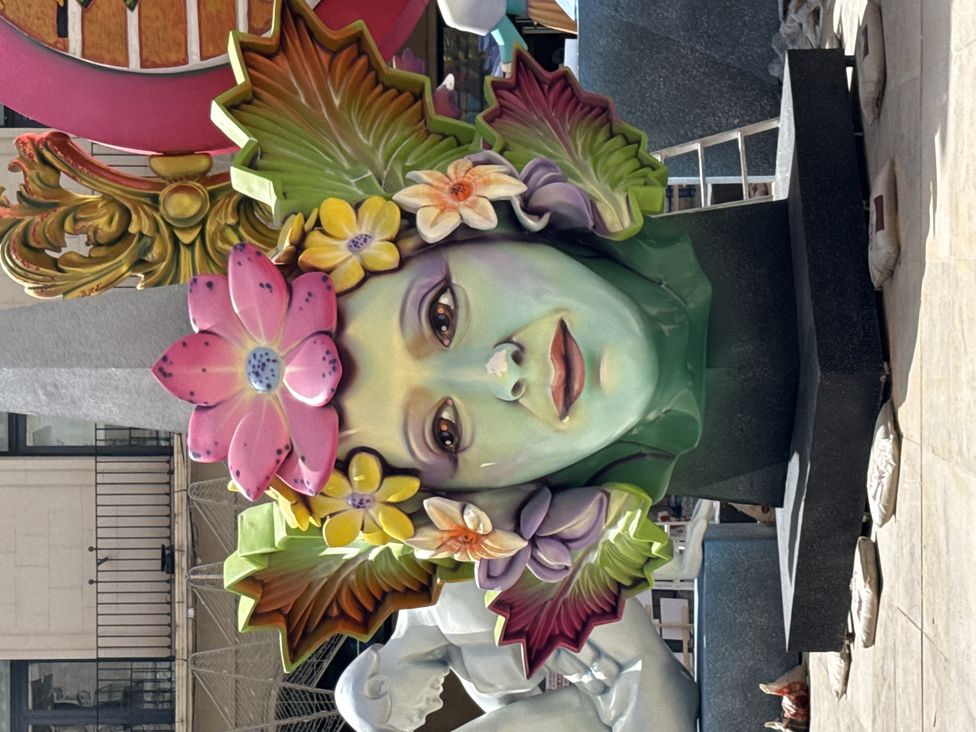 The media file displays a close-up, vertical shot of a large, colorful art installation, likely a float or a monument. The central subject is a stylized, pale green face with large, expressive eyes and rosy cheeks. The face is adorned with a vibrant headdress of oversized, sculpted flowers in shades of pink, yellow, and orange, interspersed with large, leaf-like structures in green, brown, and purple. Some of these elements appear to be made of plastic or painted material, with a glossy finish.
Beneath the face, a partially visible white sculpture of a torso and arms is depicted, suggesting the face is part of a larger figure. The background shows a bustling urban environment with hints of buildings and scaffolding, implying the installation is outdoors, possibly in a public square or street. The lighting suggests it is daytime, with bright sunlight casting distinct shadows, particularly on the darker, textured surfaces in the background, which appear to be stone or concrete.
The overall impression is festive and theatrical, potentially related to a festival or public celebration. The art piece itself evokes a sense of nature and beauty, with the floral elements and the serene, almost ethereal expression of the face. There are no discernible signs, text, or specific indications of the time of day or weather conditions beyond the presence of sunlight. The image is oriented vertically, and the composition focuses tightly on the intricate details of the art piece.