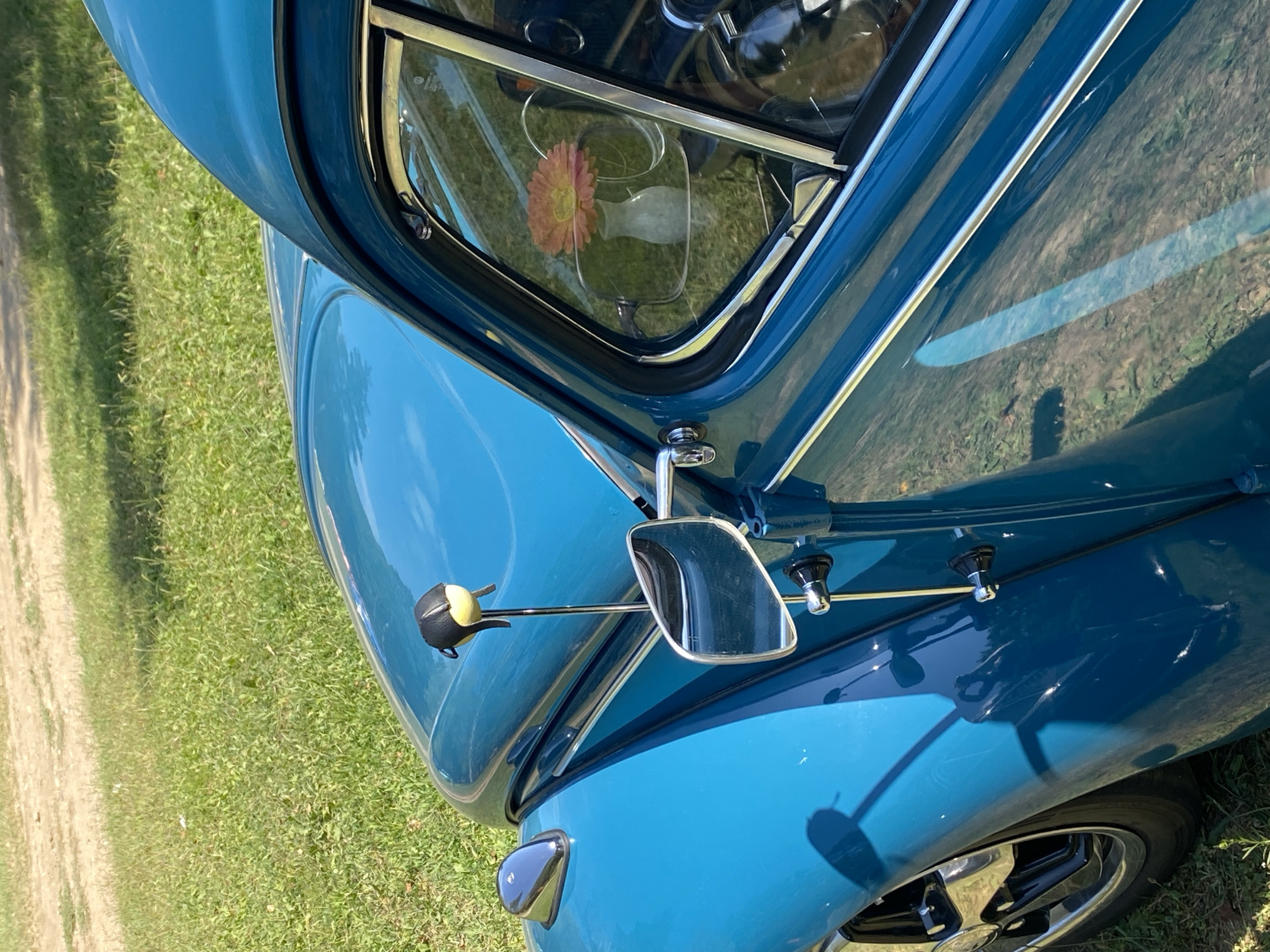 A close-up, eye-level shot focuses on the driver's side of a vintage, teal-colored car parked outdoors on a grassy area. The car's polished surface reflects the surrounding environment, including greenery and a dirt path. A chrome side mirror is attached to the door, and a decorative black and yellow ornament is mounted on a slender rod above it. Inside the car, visible through the window, is a small white vase holding a single pink and orange daisy. The car's front fender and a portion of its wheel, featuring a chrome rim and black tire, are also visible on the left side of the frame. The scene appears to be set in Bezymyannoe, Russia, with the overall impression being one of a well-maintained classic vehicle on a sunny day.