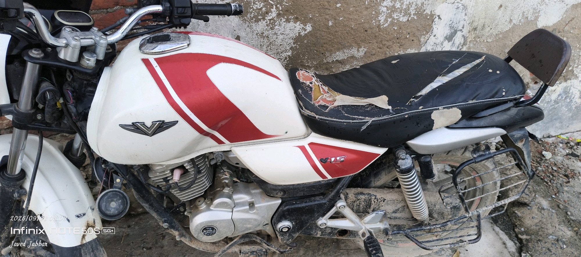 The image shows a white and red motorcycle, specifically a Bajaj V15, parked outdoors on a dirt and concrete surface, likely a street or alley. The motorcycle is the primary subject. It appears to be in a state of disrepair or abandonment, with a torn seat, visible dirt and mud on the tires and frame, and possibly some rust. The background consists of a weathered, peeling wall. The motorcycle has a chrome handlebar, a white fuel tank with red decals, a black seat with significant damage, a silver rear fender, and a black metal luggage rack over the rear wheel which also has a protective grille. The front of the motorcycle is visible, including the front suspension, a white fender, and a round horn. The image is timestamped "2026/09/08 08:09" and shows text overlays "Infinix NOTE 505" and "Javed Jabbar." The scene is located in Bahera, India, as indicated by the context provided. There are no people visible in the image.