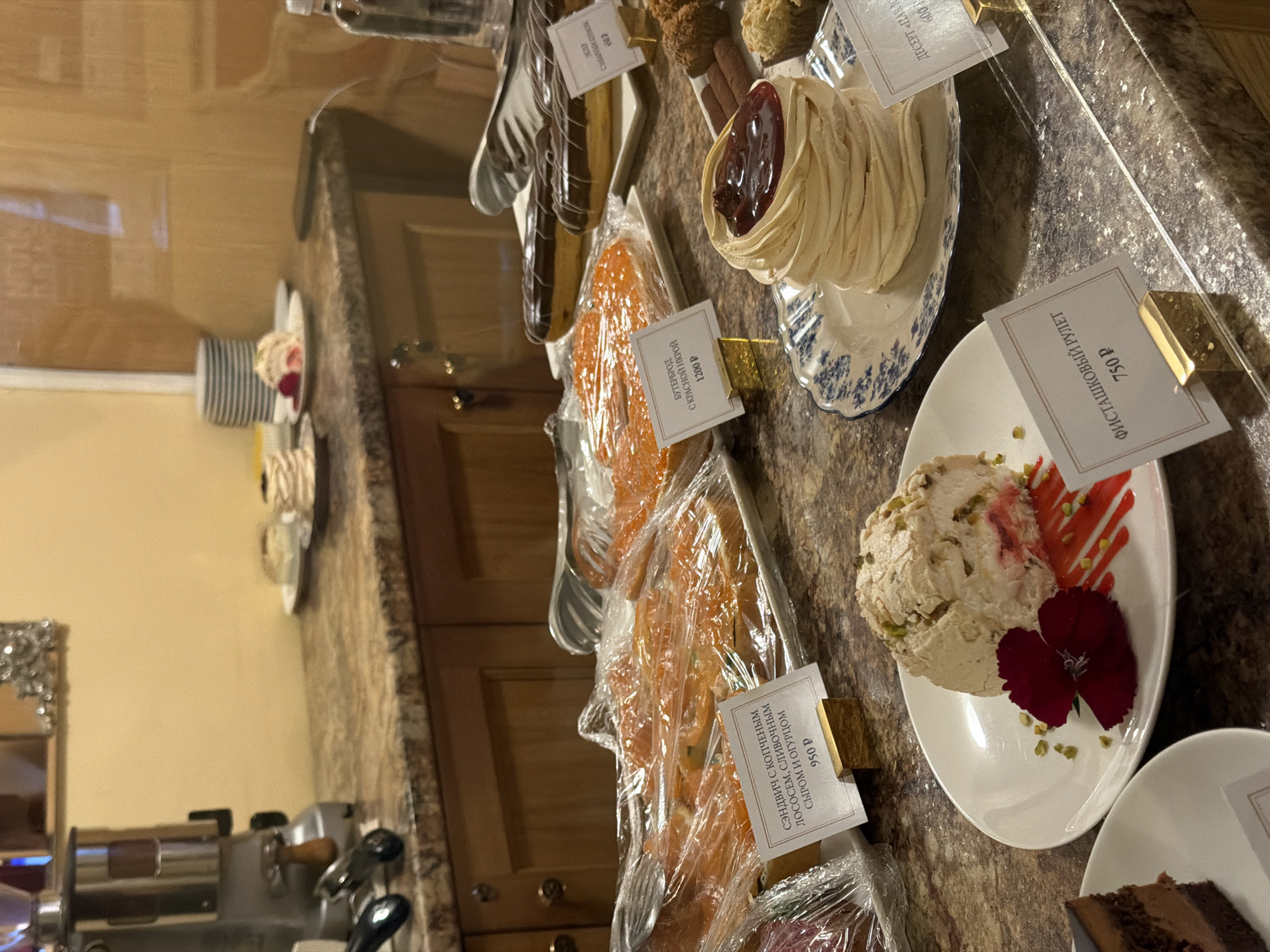 This image depicts a display of desserts in what appears to be a cafe or bakery, possibly located in Lednevo, Russia. The scene is a close-up shot focusing on a counter filled with various sweet and savory items. In the foreground, to the left, is a slice of chocolate cake on a white plate. Next to it, a larger white plate holds a meringue-based dessert topped with chopped pistachios and garnished with a dark red flower and a red drizzle. A small white card with text is placed in front of this dessert.
Behind this, on a blue and white patterned plate, sits a delicate, stacked meringue dessert with a dark red berry topping. Another white card is visible in front of this item. Further back, a tray of what appears to be salmon sandwiches, covered in plastic wrap, is displayed with a white card indicating the price. Adjacent to this tray is another dish covered in plastic wrap, containing what looks like a large, flat orange-colored pastry or cake. A white card with Cyrillic text and a price is also present. To the right, several chocolate éclairs are arranged on a white platter, with a price card in front. In the background, a coffee machine is visible on the counter. The overall environment suggests a commercial food establishment.