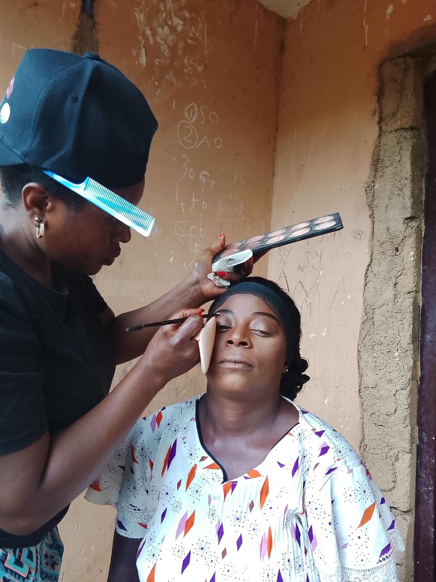 A makeup artist is applying makeup to a woman's face in what appears to be an indoor or semi-outdoor setting. The makeup artist, wearing a black cap and a black shirt, holds a palette of eyeshadows in her right hand and uses a makeup brush in her left hand to apply makeup to the woman's eyebrow. The woman, wearing a white patterned shirt and a black headband, has her eyes closed and appears to be relaxed. The background is a wall with some writing on it and what appears to be a rough, possibly unfinished, building material. The location is in the city of Unknown, Unknown.