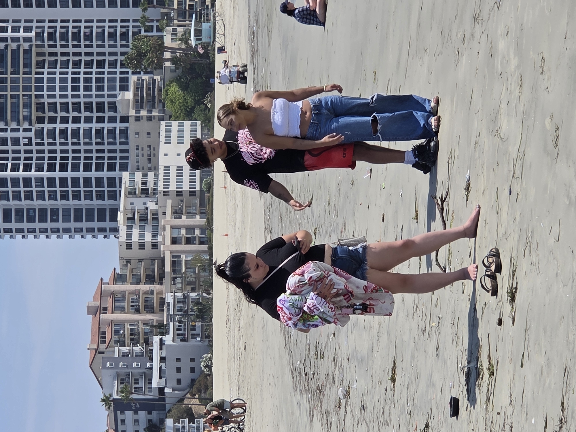 On a sandy beach, likely in Long Beach, California, three young adults are visible. A woman in a black top and denim shorts stands in the foreground, holding a colorful towel and looking at her phone. To her right, a couple is interacting. The man, wearing a black t-shirt and red shorts, has his arm around the woman, who is in a white tube top and ripped jeans. In the background, other beachgoers and buildings are visible, including a tall, modern high-rise and lower-rise apartment buildings. The beach appears somewhat littered with debris.