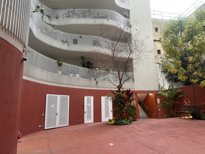 This image depicts a modern architectural scene within a courtyard in Tel Aviv-Yafo, Israel. The primary subject is a multi-story building featuring a prominent, deeply curved, light-colored (off-white/beige) facade. Each of its visible four levels incorporates a balcony lined with white, closely spaced vertical railings. Potted plants are visible on these balconies, with bicycles stored on one upper balcony.
At ground level, a reddish-orange paved courtyard area leads to a curved wall of the same color. Along this wall, three white louvered doors are present: one double door centrally located, and two single doors to its right, appearing to be utility or storage entrances. A red fire hydrant cover is visible on the ground near the left side of the double door. A small, dark security camera is mounted on the curved reddish-orange wall on the left.
To the right, a reddish-orange painted stairwell entrance leads downwards, marked by a green "EXIT" sign. The courtyard features distinct vegetation: a bare-branched deciduous tree stands in the foreground, while a cluster of vibrant green, leafy trees occupies the right side, partially obscuring an older, taller, light brown building with small, square windows in the background. Lush green foliage, including tropical plants and shrubs, is cultivated around the base of the bare tree and near the stairwell. A dark-stained wooden slatted bench with a metal frame is positioned in the right portion of the courtyard. No individuals are present, and no actions or interactions are depicted.