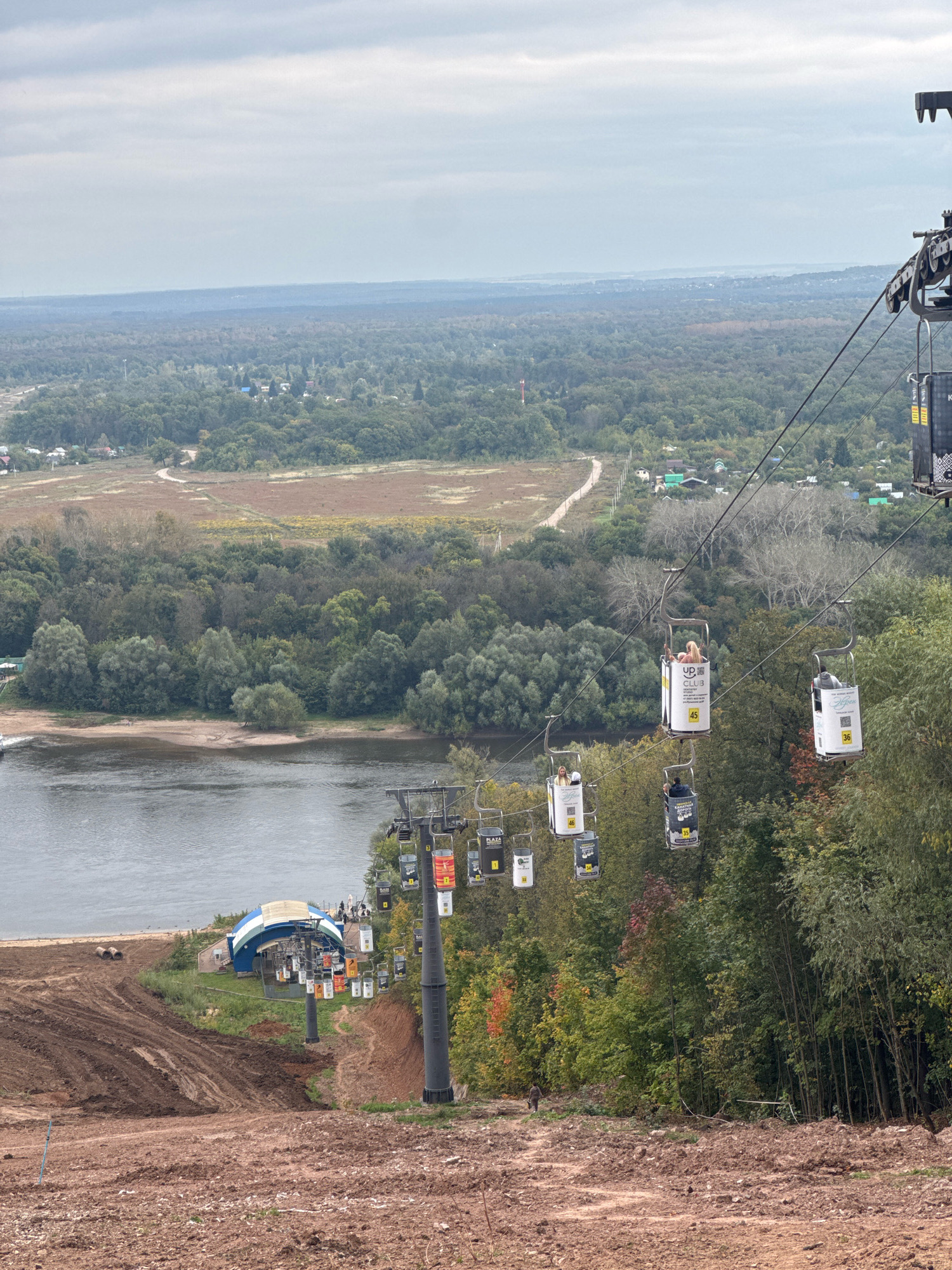 This image depicts a landscape featuring a ski lift in operation, set against a backdrop of forested hills and a river. The ski lift, with multiple open-air cabins, is suspended from cables stretching across the frame. Some cabins are occupied by passengers. The foreground shows a muddy, excavated slope, suggesting recent construction or preparation for a winter sport. A riverbank with a small building and several cylindrical objects is visible below the slope. In the distance, a vast expanse of trees stretches towards a hazy horizon, with scattered houses and clearings indicating a semi-rural or suburban environment. The sky is overcast, with a soft, diffused light. Based on the presence of a ski lift and its design, the scene is identified as being at the "Aksakovo" ski resort located in Ufa, Russia.