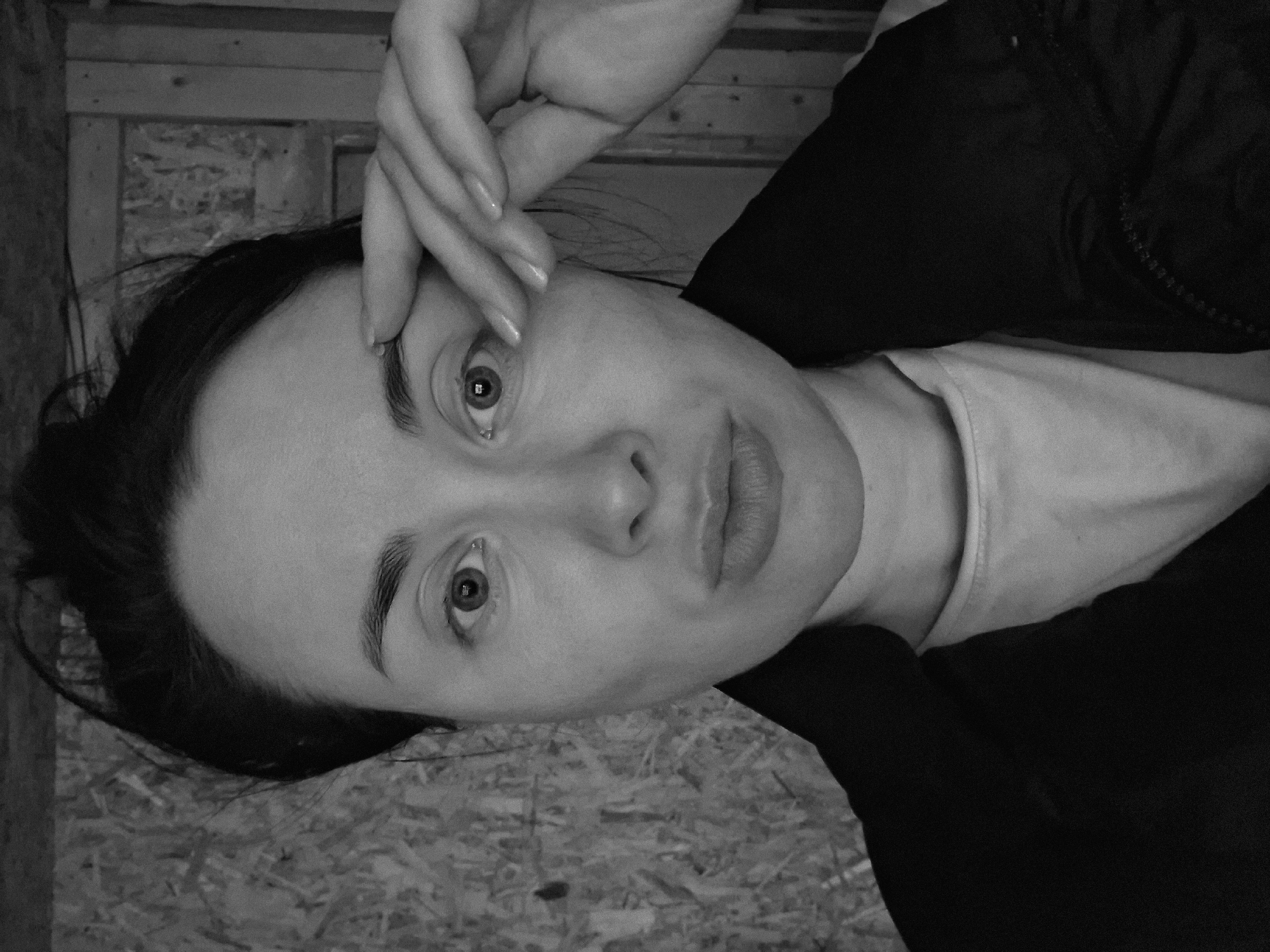 A black and white, medium-close up shot depicts a woman indoors. Her face is centered, with her right hand placed near her forehead. She is wearing a light-colored shirt under a dark jacket. The background features wooden wall panels. The location in the city is Unknown, Unknown.