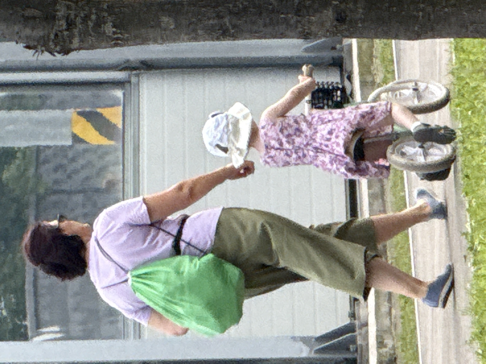 In an outdoor setting, an adult woman and a young child are walking on a paved surface. The woman, with dark hair pulled back and wearing sunglasses, has a green drawstring bag slung over her left shoulder and is wearing a light purple t-shirt and olive green pants. The child, facing away from the camera, is wearing a pink floral dress and a white hat with a brim. The child is seated on a small bicycle with two wheels, holding onto the handlebars. The woman's right hand is extended towards the child, suggesting they are holding hands or the woman is guiding the child. The background includes a building with a textured wall and a yellow and black diagonal striped warning sign. A portion of a tree trunk is visible on the right side of the frame. The lighting suggests it is daytime.