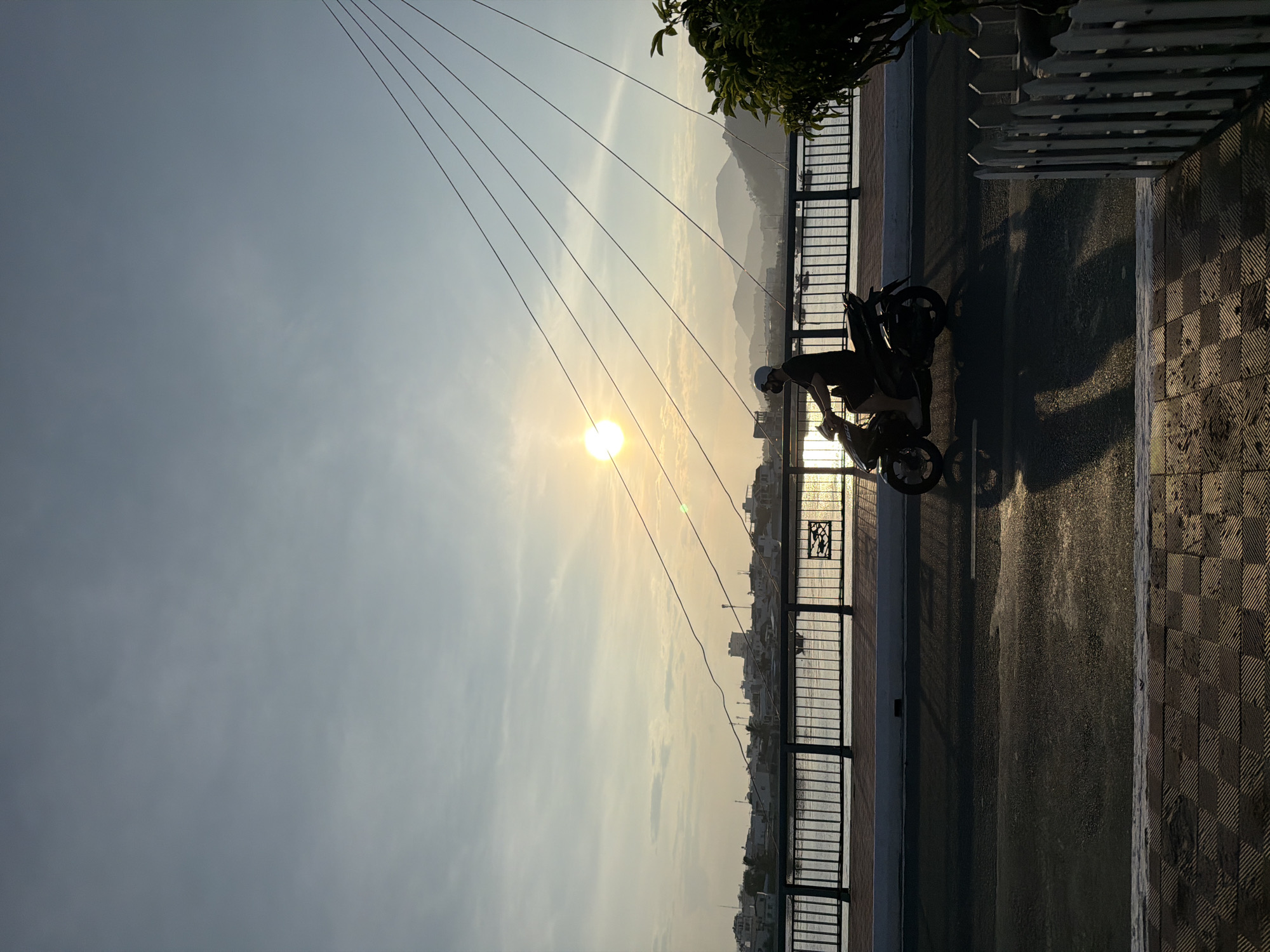 This image depicts a sunrise or sunset over a body of water in Bac Nha Trang Ward, Vietnam. The scene is illuminated by strong backlighting from a bright, low sun positioned in the upper left, casting long, dark shadows toward the viewer.
The sky transitions from a light blue-grey at the top to warmer golden-yellow and orange tones around the sun, with faint, scattered clouds. Numerous dark, tensioned cables of a cable-stayed bridge extend diagonally from the upper right portion of the frame downwards and to the left, intersecting the sun.
In the distant background, a series of layered mountains appear as hazy, progressively lighter silhouettes against the horizon. Below these, a city skyline with various buildings is silhouetted on the far bank of a wide body of water, which reflects the golden light. Several small boats are visible on the water's surface.
A dark metal bridge railing, composed of vertical bars and a decorative panel featuring cut-out fish designs on its right side, horizontally traverses the midground, separating the roadway from the water below.
The immediate foreground features a dark, reflective asphalt road surface with a faint white line. Elongated shadows from the main subject stretch across this road. To the left of the road, a sidewalk is paved with dark, textured, checkered tiles. On the far right, a white picket fence and dark green foliage are partially visible at the bottom edge.
A lone individual, wearing a light-colored helmet and a dark short-sleeved top, is riding a dark-colored scooter towards the right side of the frame. The person and the scooter are largely silhouetted, but the number "150" is faintly discernible on the scooter's side.