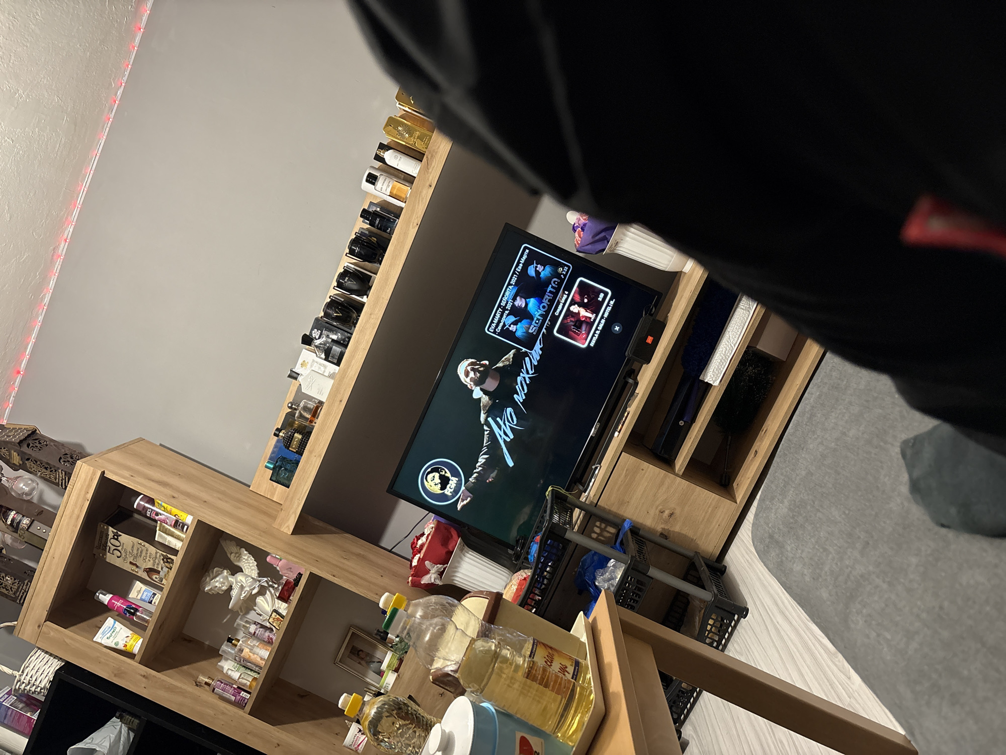 The image shows an indoor scene, possibly a living room, with a light grey wall adorned with red LED lights. A wooden shelving unit displays various items, including perfumes, bottles of cooking oil, decorative items, and toiletries. A television screen shows a video playing, displaying text in Cyrillic and Latin script, along with song titles like "SENORITA" and "Ако може". A part of a person's body, clad in black clothing, is also visible, possibly indicating the person taking the picture. The image may be located in Kazichene, Bulgaria, given the Cyrillic script present on the TV screen.
