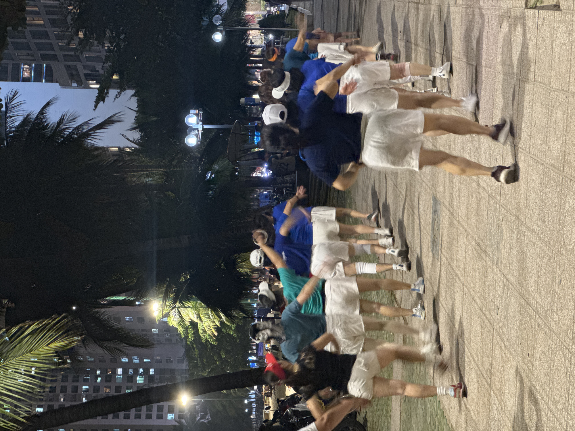 A long exposure shot captures a line of people exercising along a paved walkway in what appears to be a park or public space. The location appears to be in a city at night, evidenced by illuminated buildings in the background and streetlights.
The people in the line are wearing a combination of dark-colored shirts and white shorts. Some are wearing hats, including baseball caps. The long exposure has created a blurred effect, indicating movement during the shot.
Along the side of the walkway, palm trees are visible, adding to the location context. Further off, there are buildings with lit windows.
The foreground pavement has a textured pattern, possibly made of pavers.