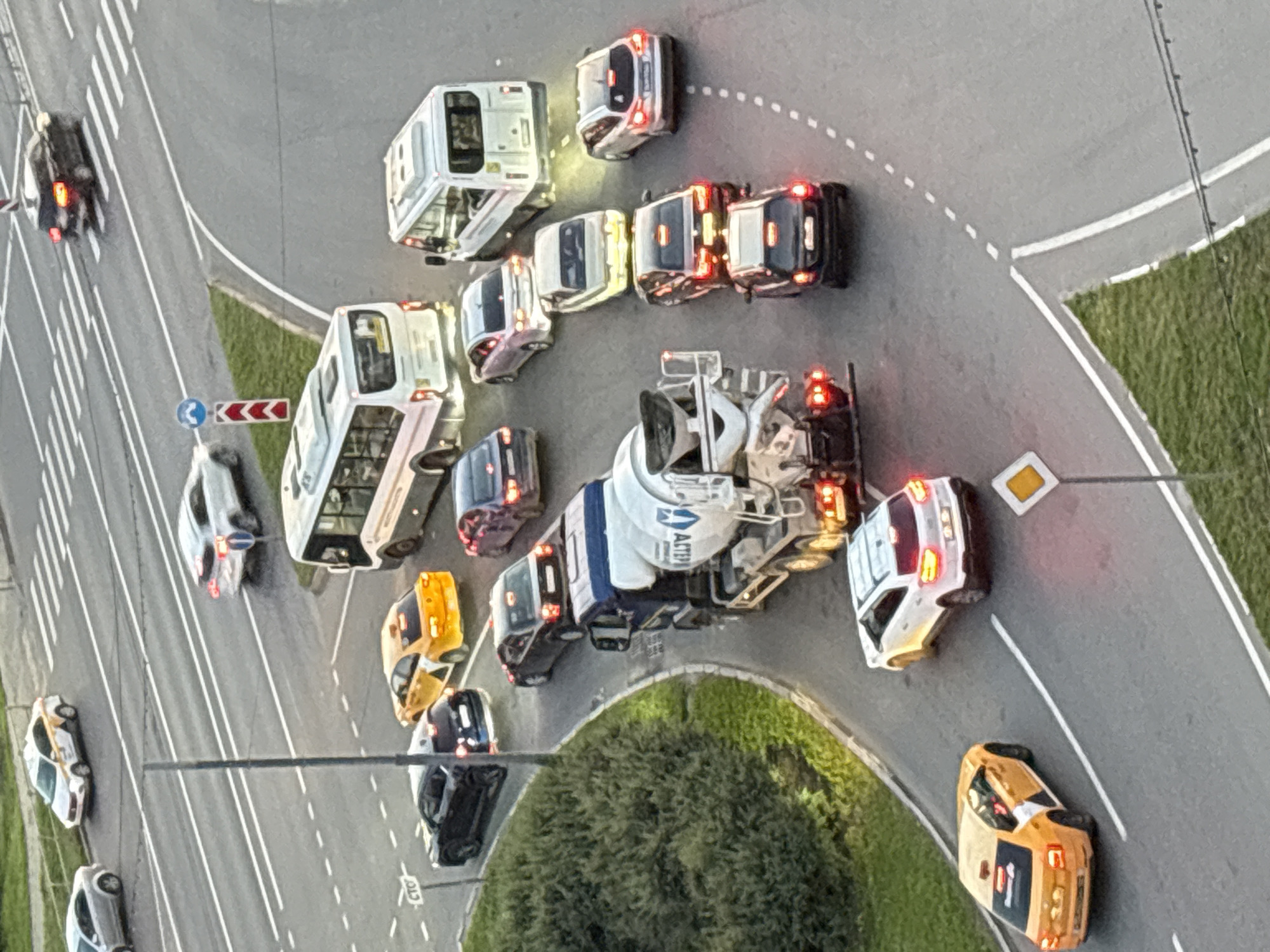 This media file is an aerial, slightly angled view of a traffic scene, likely during daylight given the illumination. The scene appears to be a road intersection or a roundabout, with multiple vehicles present.
The subjects are various vehicles, including a concrete mixer truck, several passenger cars (some yellow, some white, and at least one dark colored), and two buses. Some of the vehicles have their brake lights or hazard lights illuminated, suggesting they are stopped or moving slowly. The concrete mixer truck is prominent in the center-right of the frame. One of the yellow cars has visible text and logos on its side, possibly advertising. One of the buses appears to have text on its side as well. There are no visible people in the scene.
The activity taking place is traffic congestion. The vehicles are positioned on the road in a way that suggests they are waiting or moving through a busy junction. The road itself is asphalt, with white lane markings and a grassy verge visible along the edges. There is a traffic sign visible with a blue circle and a white arrow, as well as a red and white chevron sign. Another diamond-shaped sign with a yellow center is also present.
Notable details include the diverse range of vehicles and the implied standstill or slow movement of traffic. The lighting suggests it is daytime, and there are no immediate cues about the weather. The location is unclear, but it appears to be an urban or suburban road environment.