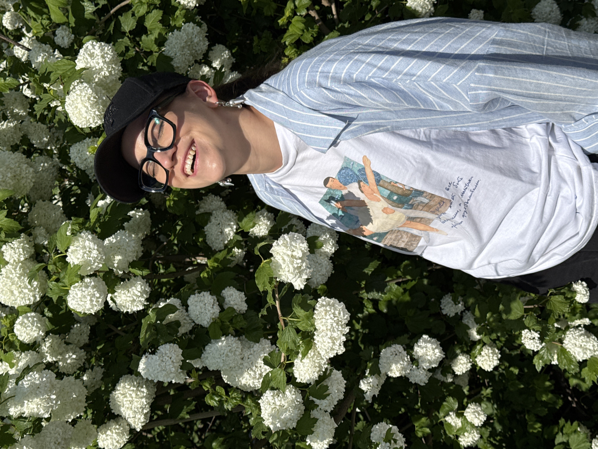The image shows a woman lying amongst a bush of white hydrangea flowers. She is smiling and wearing a black baseball cap, eyeglasses, a white t-shirt with an image printed on it, and a light blue and white striped shirt. The photo was taken outdoors in daylight. The location is Kazan, Russia.
