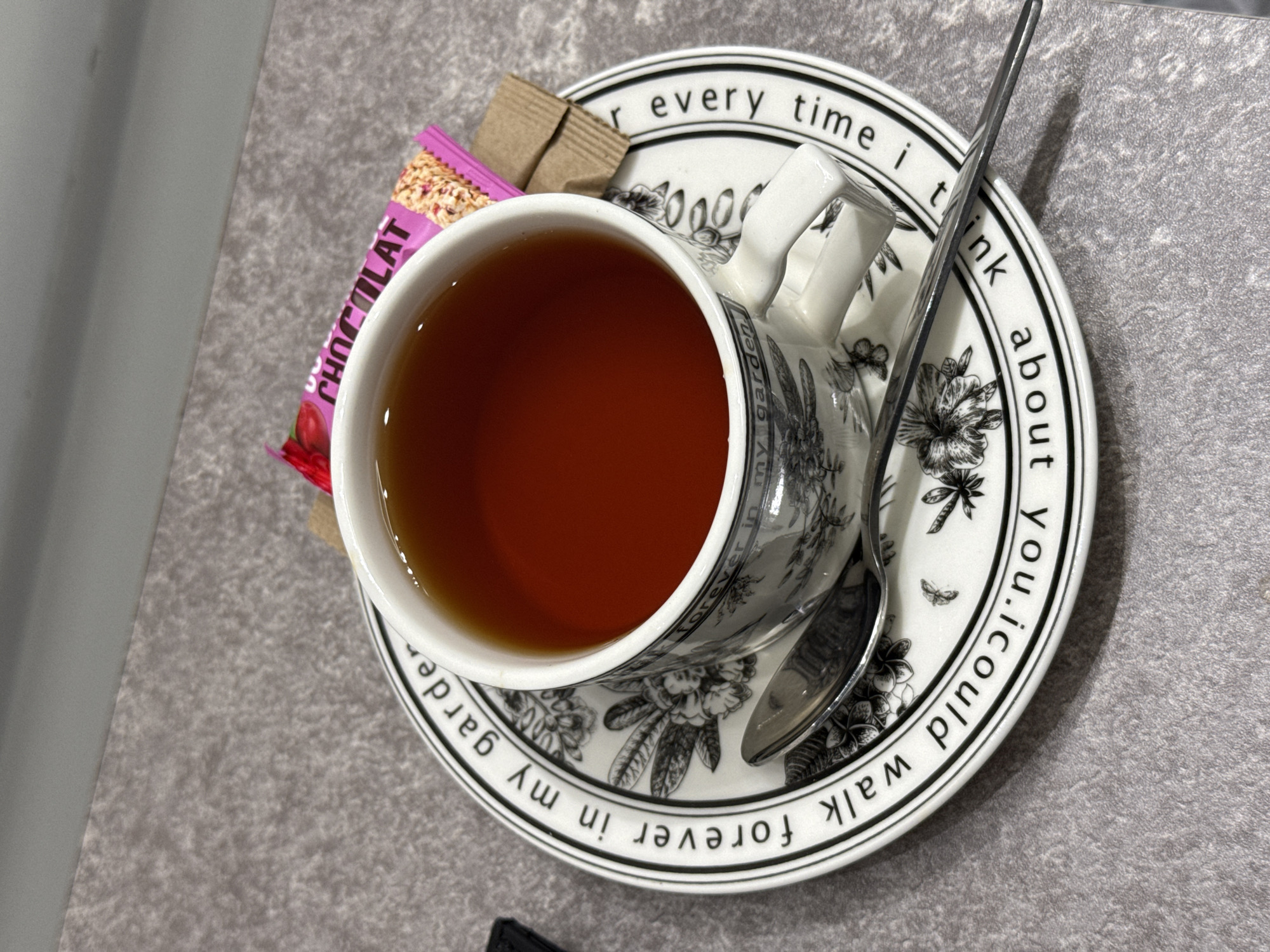 The image presents a top-down, close-up view of a tea setting. A cup filled with tea sits on a saucer, accompanied by a spoon, a small granola bar with the word "CHOCOLAT" visible on the wrapper, and two brown sugar packets. The saucer is decorated with floral designs and a black-and-white border containing a quote: "for every time I think about you, I could walk forever in my garden." The setting is placed on a grey surface. The location in Saint Petersburg, Russia, cannot be determined from the information presented in the image.