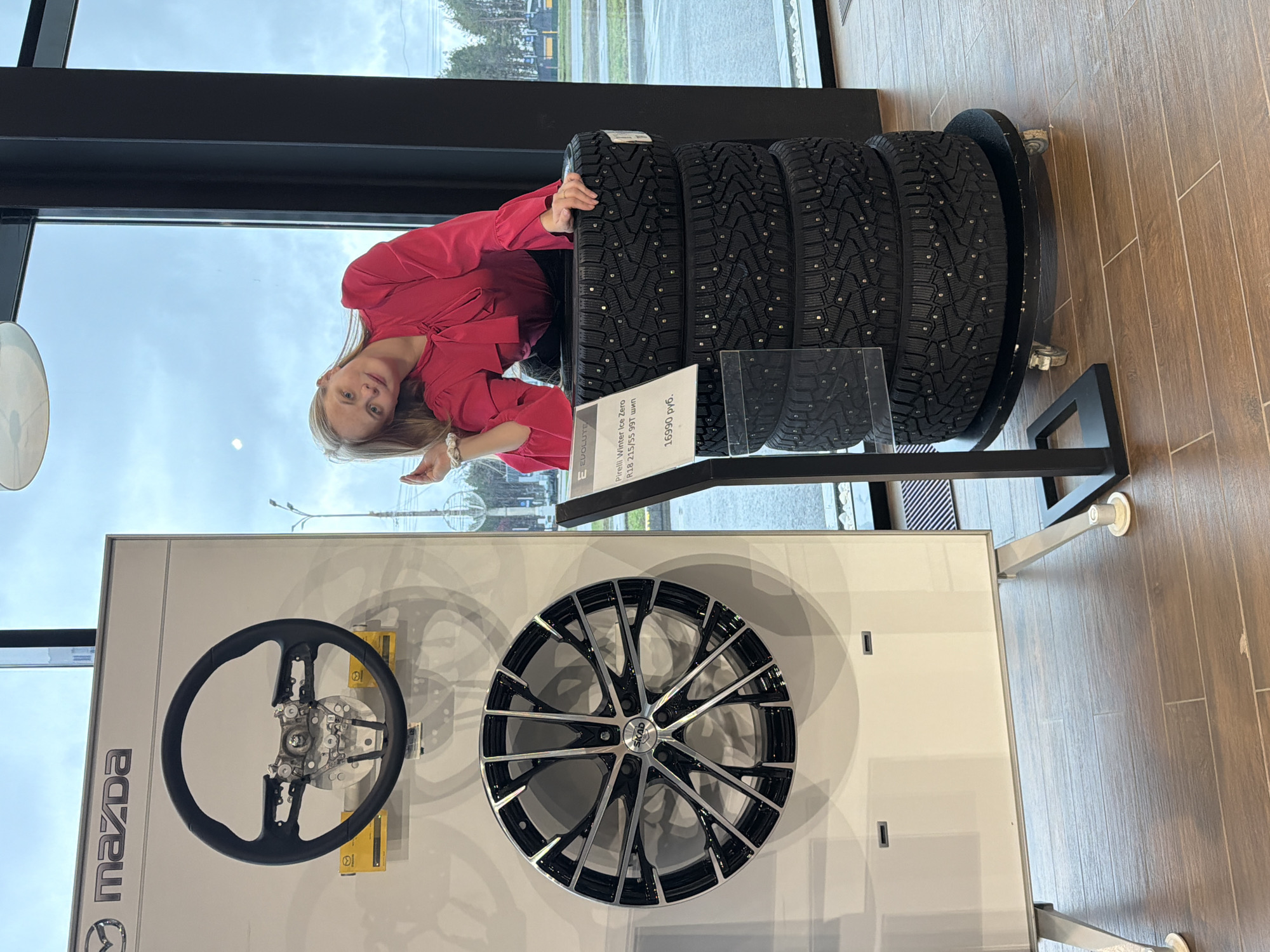 The photograph was taken indoors in Izhevsk, Russia. The picture features a blonde woman in a red blouse leaning against a stack of four studded Pirelli Winter Ice Zero tires on a rolling platform. The tires are priced at 16990 RUB. Behind the woman and the tires is a window showing an overcast outdoor scene. In the foreground, below the tires, is a display showcasing a Mazda steering wheel and a Skad rim. The Mazda logo is visible on the left side of the display. The background is blurred, suggesting a showroom environment. The floor is wooden. The woman is making a hand gesture.