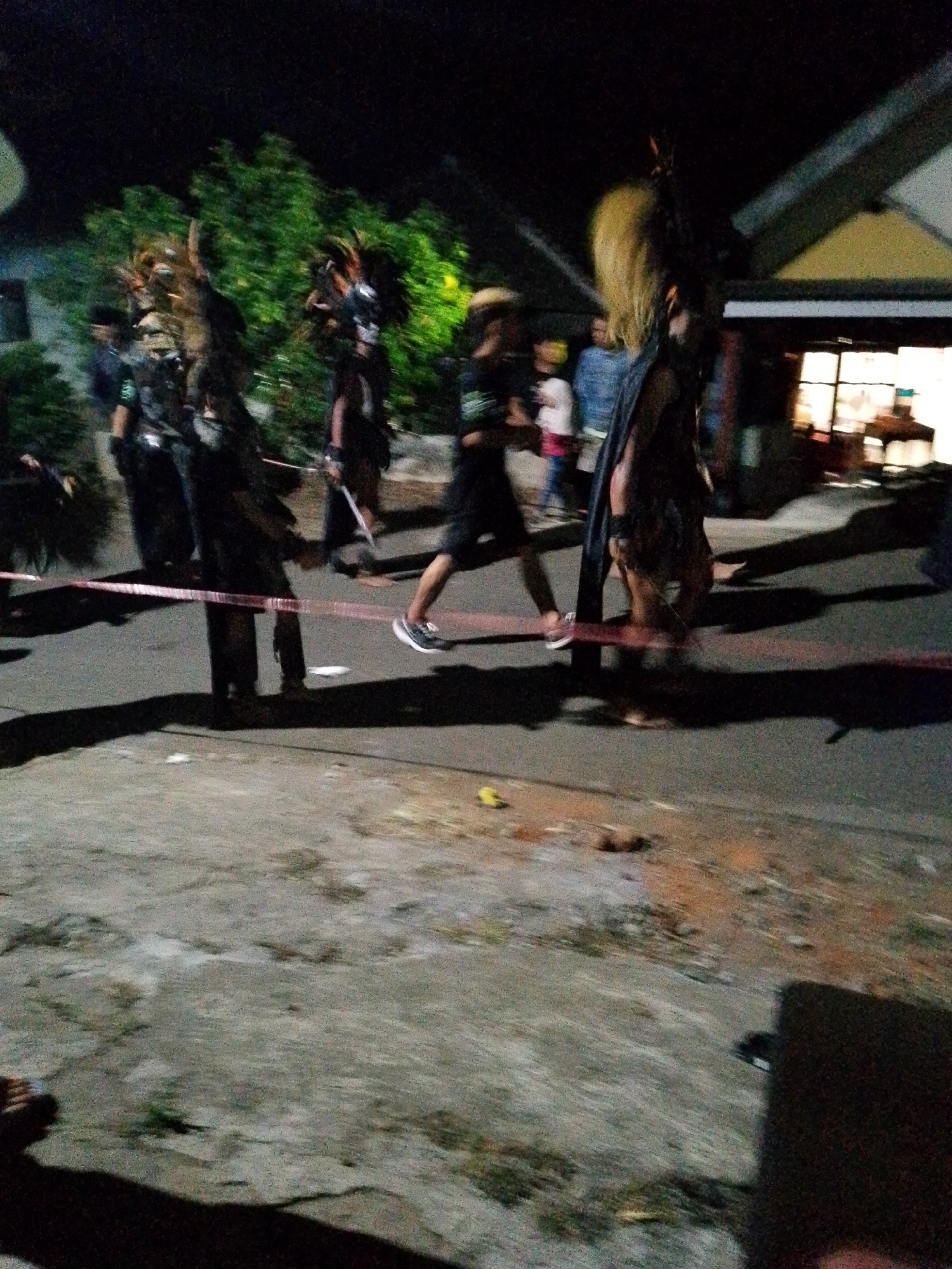 This is a blurry, nighttime photograph taken in Babadan, Indonesia, showing a group of people participating in a cultural performance. Several individuals are wearing elaborate headdresses with what appear to be feathers or animal parts, and some are adorned with dark, patterned clothing. One person in the foreground, on the right side of the image, is wearing dark shorts and sneakers and is in motion, suggesting a dance or procession. A red and white ribbon or tape is stretched across the street in the mid-ground, possibly marking a performance area or barrier. In the background, a building with illuminated windows suggests a residential or commercial area, and lush green trees are visible to the left. The lighting is dim, and the motion blur indicates the photo was likely taken with a long exposure or by someone moving.