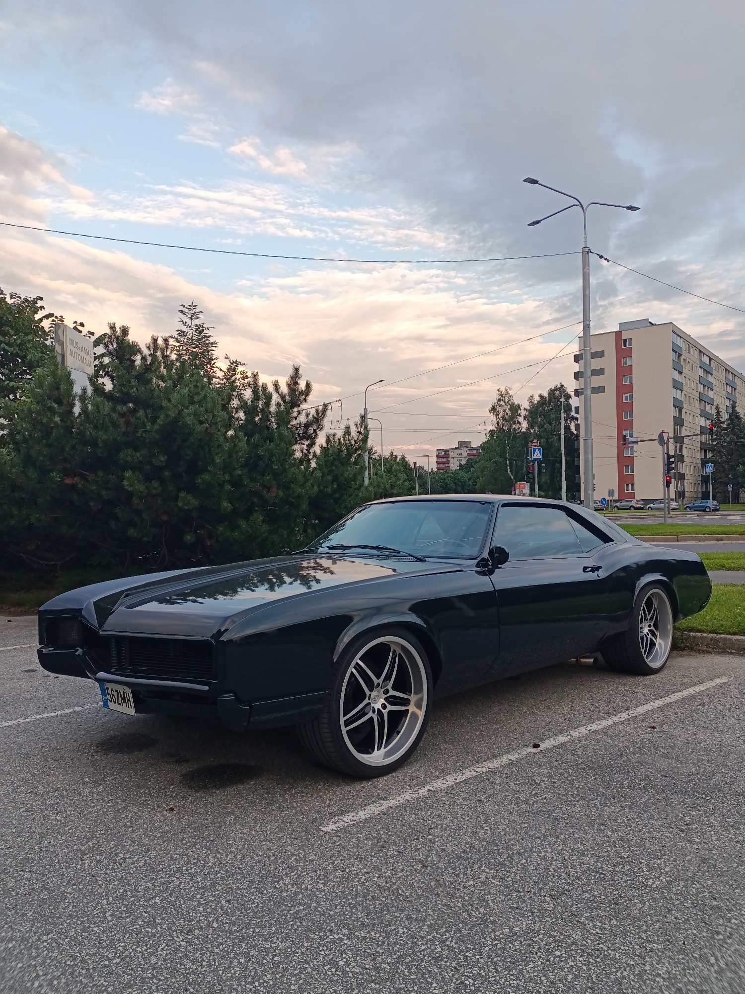 A black vintage Buick Riviera is parked in a lot at the Mustamäe Autokoda, located at Kadaka tee 48, Tallinn, Estonia. The car is angled towards the viewer, with its front left wheel turned out. The vehicle has a sleek, black exterior and custom silver rims. Behind the car, a row of evergreen trees forms a dense hedge. In the background, there are apartment buildings, streetlights, traffic lights, and a street sign with the inscription "MUSTAMÄE AUTOKODA". The sky is partly cloudy with a blend of blue and grey hues, suggesting it is either early morning or late evening. The ground is paved asphalt with visible parking lines. No people are visible in the image.