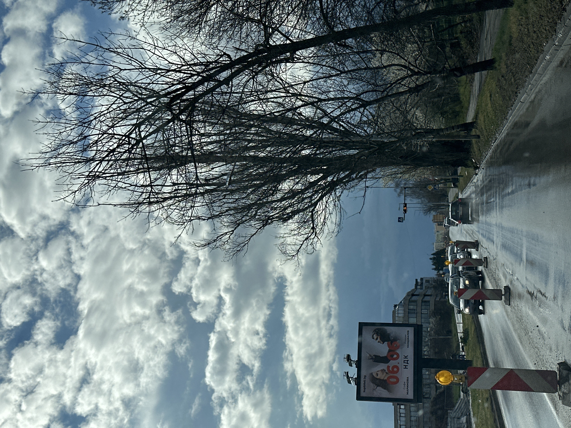 The image is taken on a street in Sofia, Bulgaria. The road is wet, possibly from recent rain, and lined with bare trees on one side. There is a line of cars stopped in traffic, likely due to road construction. Construction barriers with flashing yellow lights are visible along the road. A large billboard with an advertisement is situated near the roadside. Buildings are seen in the background. The sky is filled with streaky white clouds.