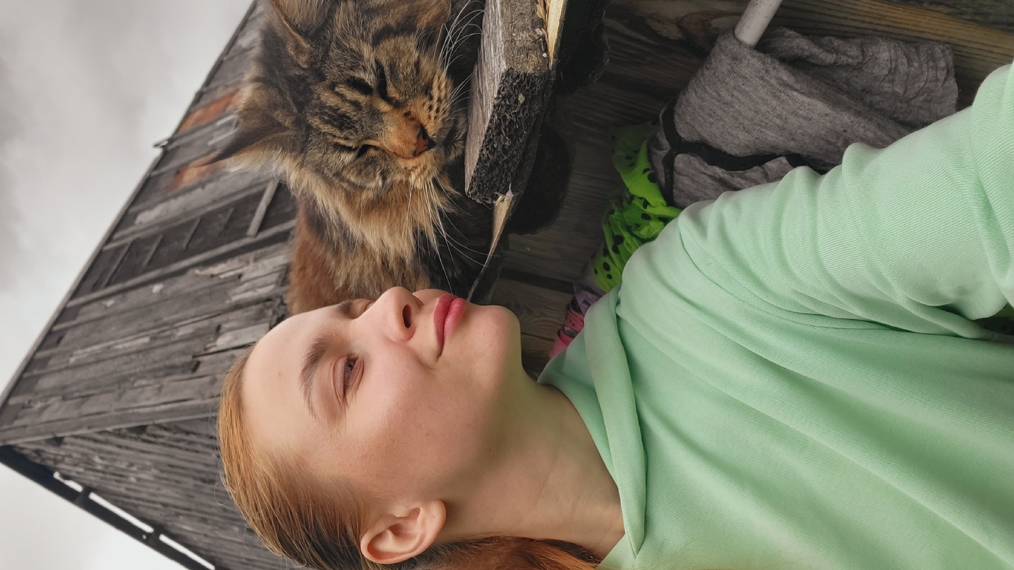 The image, captured in Зипуново, Russia, displays a close-up outdoor scene featuring a female and a large domestic cat. The female, a young adult with reddish-blonde hair pulled back, occupies the left side of the frame, presented in profile. She wears a light mint green hooded sweatshirt, and her eyes are partially closed in a relaxed expression, with a faint smile directed towards the cat.
Adjacent to her, on the right side of the frame, a large, long-haired tabby cat, consistent with a Maine Coon breed, rests its head on a rough, dark wooden plank. The cat's eyes are closed, indicating a state of rest or sleep. The background reveals a weathered, dark wooden building constructed from vertical planks, with an overcast sky visible above its structure. Below the cat and the supporting plank, a green fabric with dark spots and a grey, textured fabric are visible, alongside a partial view of a white pipe or rod.
