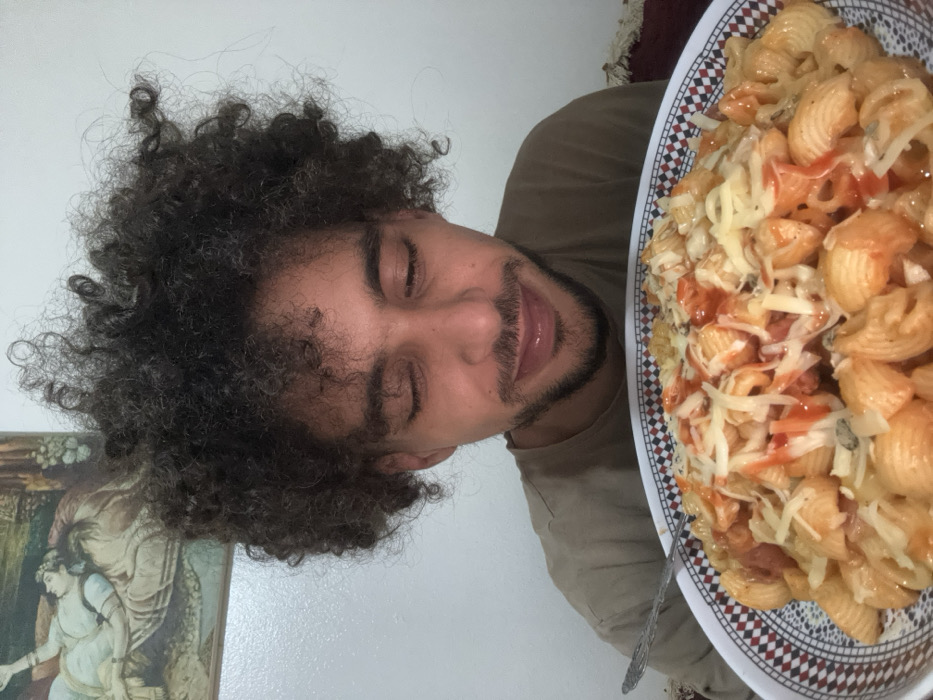 A man with curly dark hair and a beard is pictured alongside a large plate of pasta shells. The pasta is topped with a red sauce and shredded cheese. The plate features a checkered pattern on its rim. A spoon is visible in the plate. To the left of the man is a framed picture with a woman. The background is a plain white wall. The image appears to be a selfie.