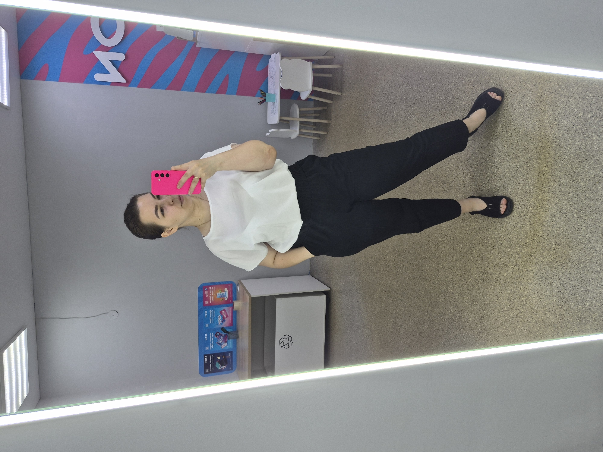 In Chaikovsky, Russia, a woman stands in front of a large mirror, taking a selfie with a bright pink smartphone. She is wearing a white, short-sleeved, loose-fitting blouse and black, high-waisted pants. Her left hand is in her pocket. She has dark hair pulled back and is wearing black open-toed sandals. The background includes a gray wall, a recycling bin, a small table with chairs, and a brightly colored wall graphic with the word "ИСК" visible. The scene appears to be in a public space, possibly a children's play area or a retail store.