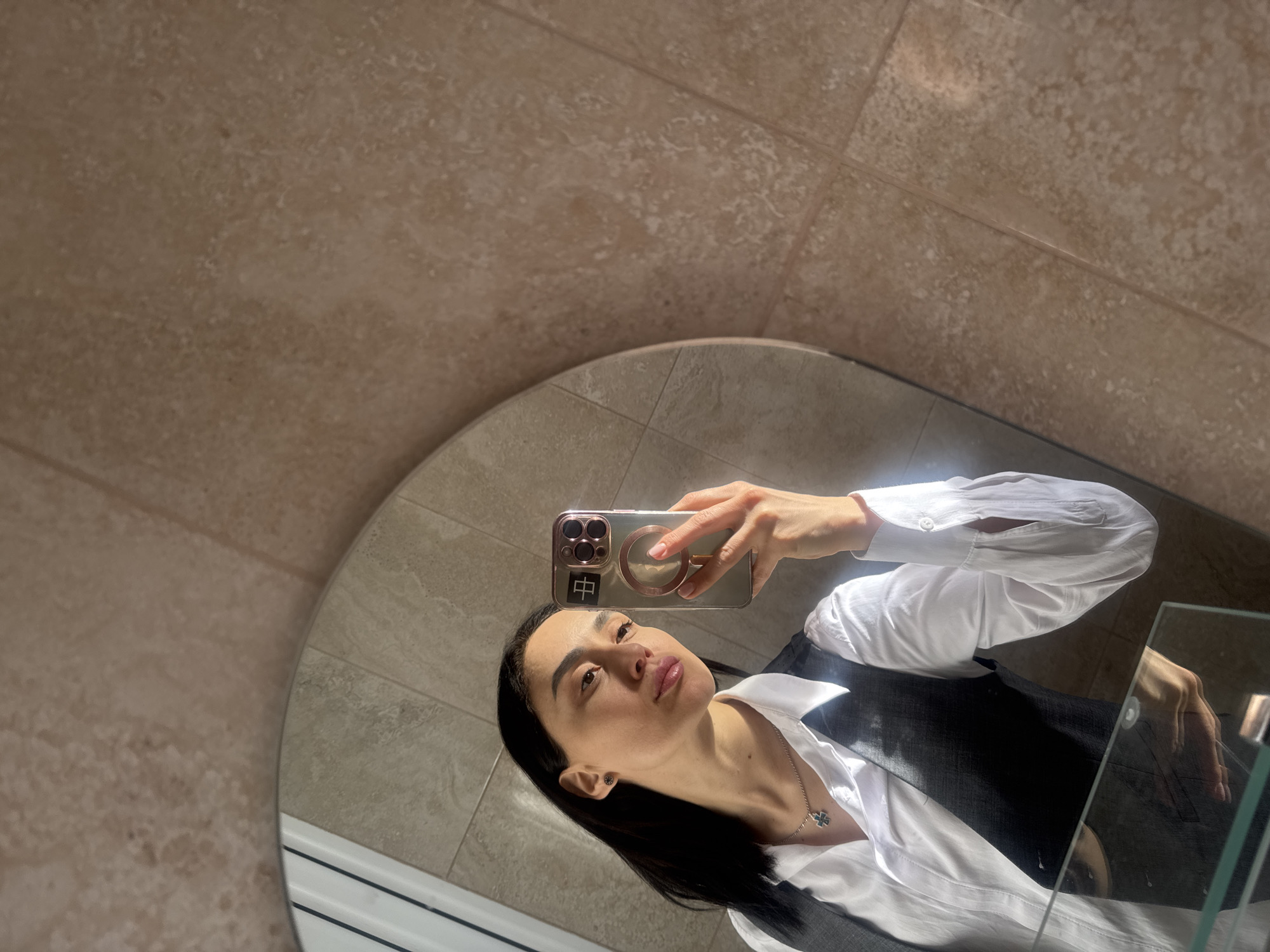 A person with dark hair is reflected in a round mirror. The person is holding a smartphone with a clear case, and the phone's reflection is also visible in the mirror. The person is wearing a white button-down shirt and a dark vest. A silver necklace with a pendant is visible around their neck. The background shows beige tile walls. The location is likely a bathroom or a room with tiled walls. This scene is probably not located in the city of Tbilisi, Georgia, as the geographical information is not available within the content of the image.