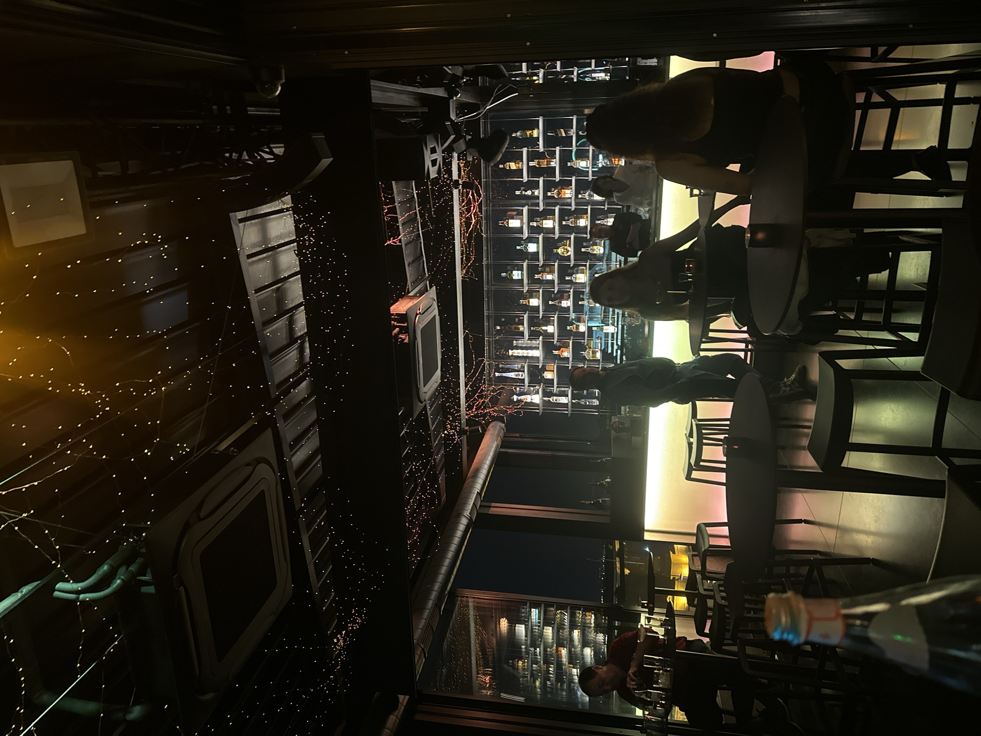 The image depicts the interior of a dimly lit bar, possibly a rooftop establishment given the view of city lights through a large window. The ceiling is adorned with numerous small, warm-toned fairy lights, creating a festive atmosphere. In the background, a well-stocked bar features shelves filled with various bottles of alcohol, illuminated from behind by soft, colorful lighting. Several individuals are present, some standing and others seated at tables and bar stools. One man in a red shirt is visible near the window, appearing to be setting down glasses. Another man stands at the bar, and further back, several people are seated at tables, their faces obscured by the low lighting and angle. The foreground is occupied by a table with a candle and a blurred bottle in the lower left corner. The overall ambiance suggests a sophisticated evening setting in an urban environment.