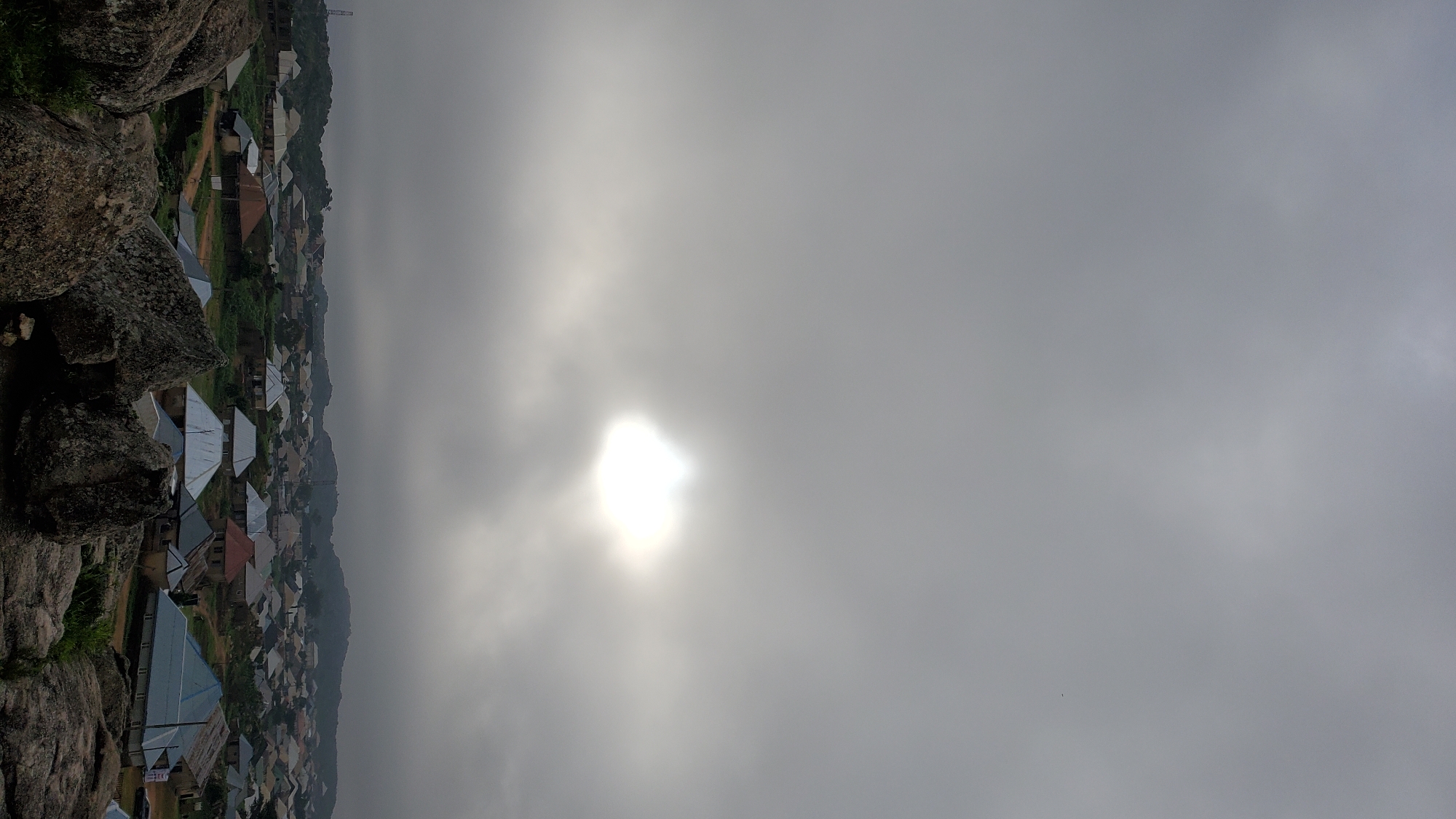 The image depicts a wide-angle, vertical view of a landscape in Jos, Nigeria, characterized by a cloudy sky and a settlement nestled among hills and rocks. The upper portion of the image is dominated by a heavily overcast sky, with a bright, diffused sun piercing through the clouds in the upper center, creating a luminous halo effect. Below the sky, a sprawling residential area is visible, composed of numerous houses with corrugated metal roofs of various colors, including silver, blue, red, and brown. These houses are clustered together, interspersed with greenery and some unpaved roads. The foreground is occupied by a large formation of rough, grey rocks, suggesting a rocky terrain. The overall atmosphere appears subdued due to the overcast conditions, with the bright sun offering a point of visual interest. No people or specific actions are discernible in the scene.