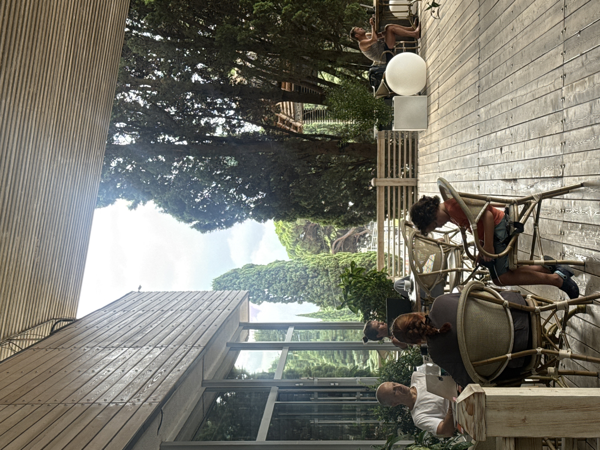 The image depicts a group of people seated at tables on a wooden deck, seemingly an outdoor cafe or restaurant area, located in Yalta, Ukraine. In the foreground, a bald man wearing a white t-shirt is seated at a table with a laptop. To his right, a woman with her back to the camera, her hair in a braid, is also seated at a table. Further to the right, a young boy in an orange shirt is seated at another table. In the background, a man in a tank top and shorts is working on a laptop at a table. The setting is lush with tall, green trees and a partially visible building with wooden siding. The overall atmosphere appears relaxed and casual.