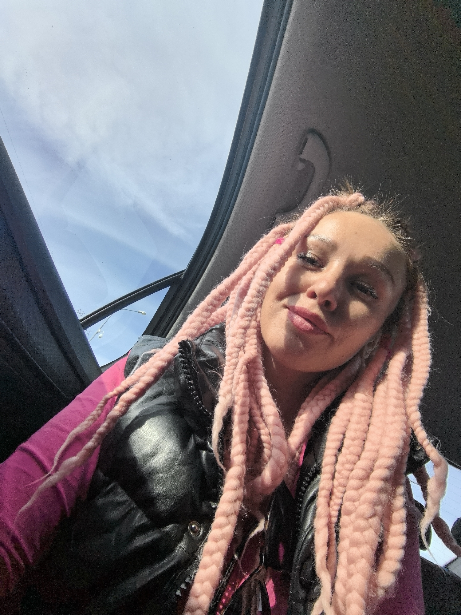 A woman with pink box braids and a black vest is sitting in a car with a sunroof. The sky is visible through the open sunroof. Based on the presence of streetlights visible through the open sunroof, this image was most likely captured in the city of Chaikovsky, Russia.