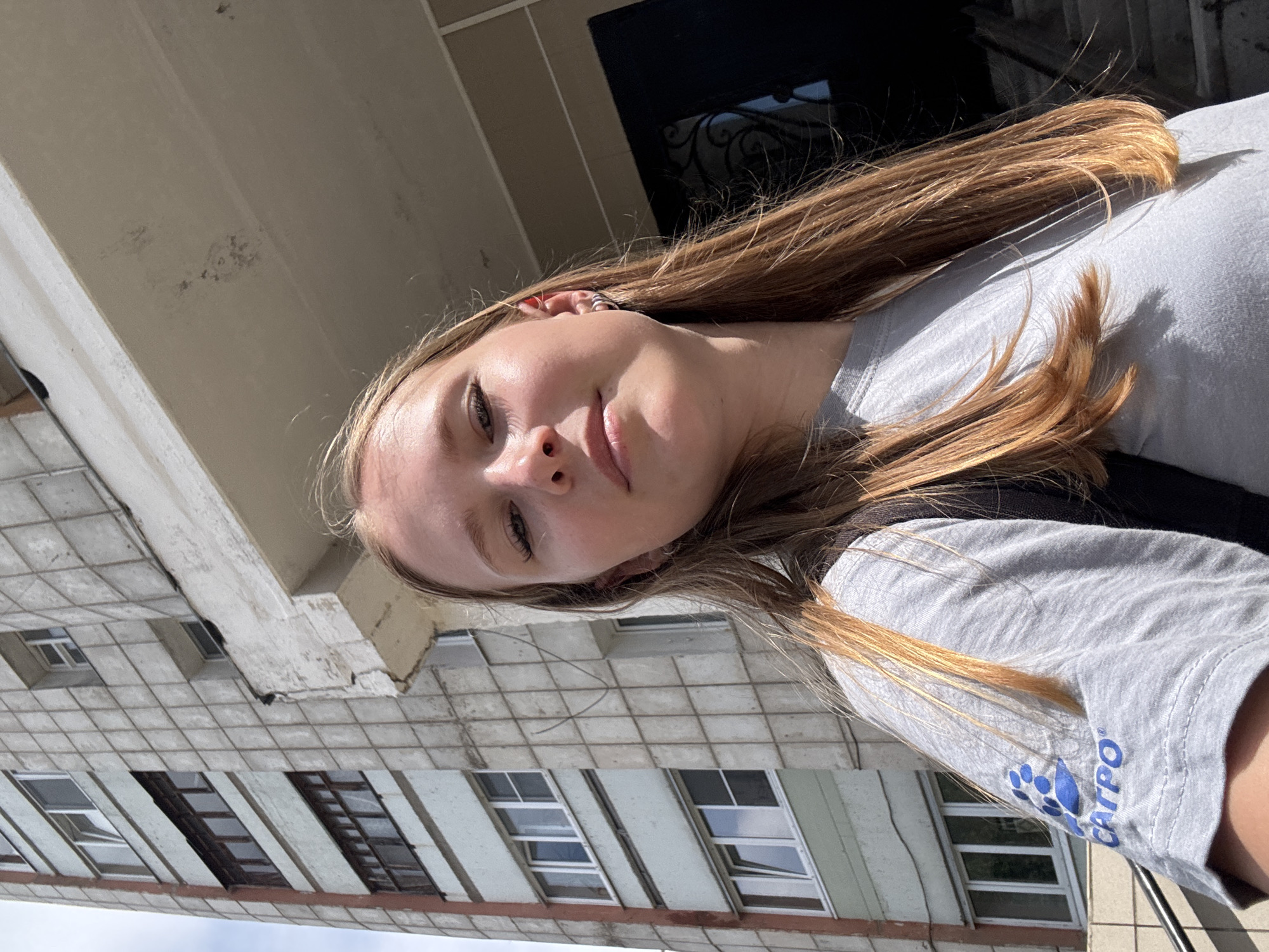 A young woman with long, blonde hair and light skin is taking a selfie outdoors. She is wearing a grey t-shirt with a blue logo on the sleeve that reads "CATPO." She has earrings in both ears, one a silver hoop and the other a red stud. Behind her, a large, multi-story apartment building with a concrete facade is visible. The building has several windows and balconies, some of which are dark. The lower portion of the building has a lighter, green-tinted concrete finish. To the right of the building, a dark doorway with ornate metalwork is visible, leading to a staircase. The sky is partly cloudy. The image appears to be taken in the city of Chaikovsky, Russia, given the architectural style of the apartment building.