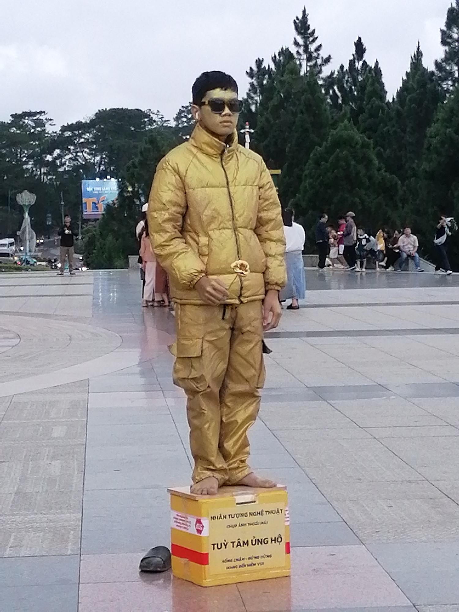 The image shows a young male dressed in a full gold outfit, including a puffer jacket and pants, standing on a yellow donation box. He is wearing sunglasses and appears to be a living statue performer. The donation box has Vietnamese text on it, indicating it is for "TUỲ TÂM ỦNG HỘ" (donations at your discretion). The background shows a crowd of people, trees, and a street with traffic, suggesting an outdoor public space. The lighting indicates it is daytime, possibly overcast. The overall scene appears to be in Da Lat, Vietnam, a city known for its French colonial architecture and lush landscapes, which is consistent with the presence of trees and a public square.